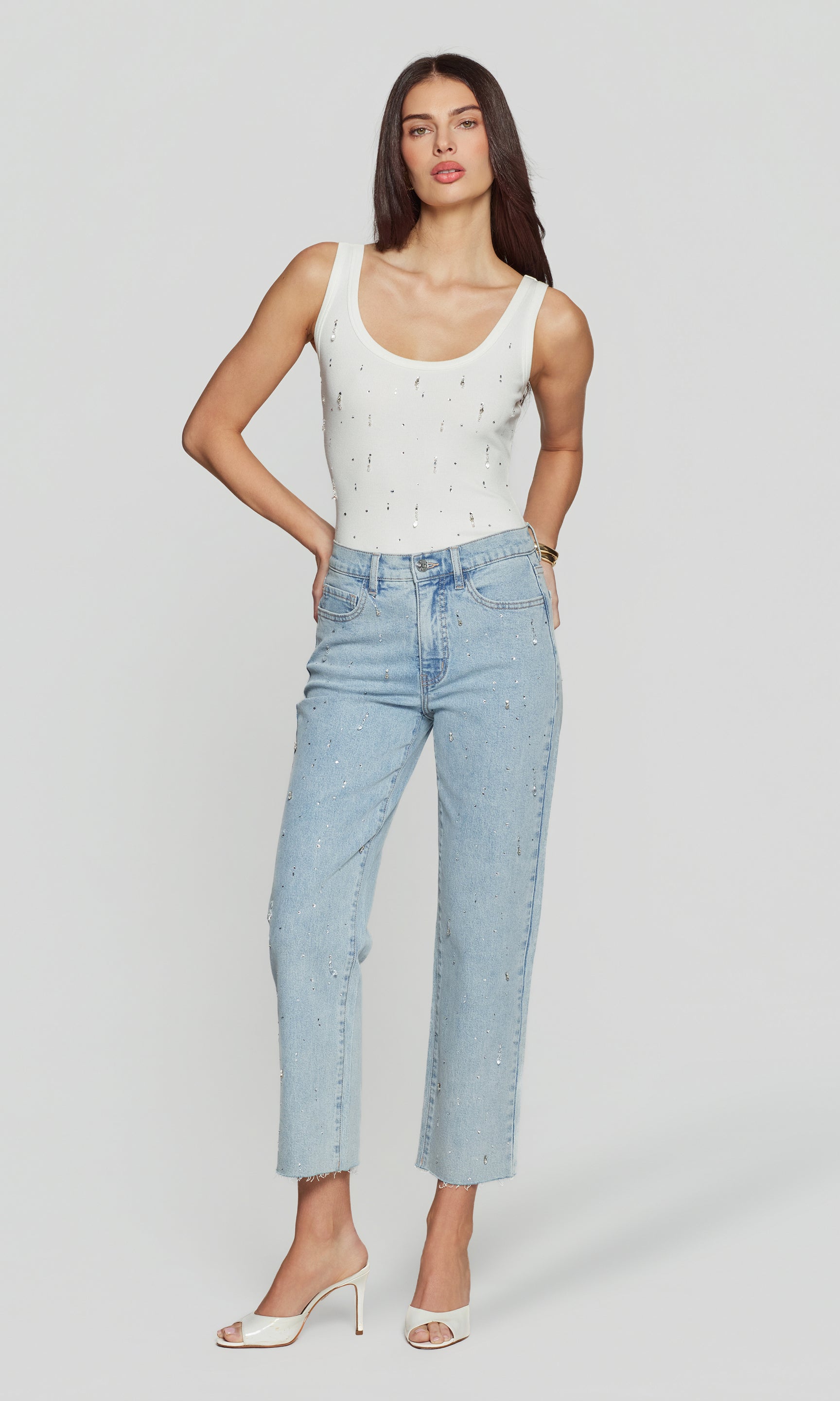 Full body image of woman wearing a white tank top and light blue jeans on a plain background