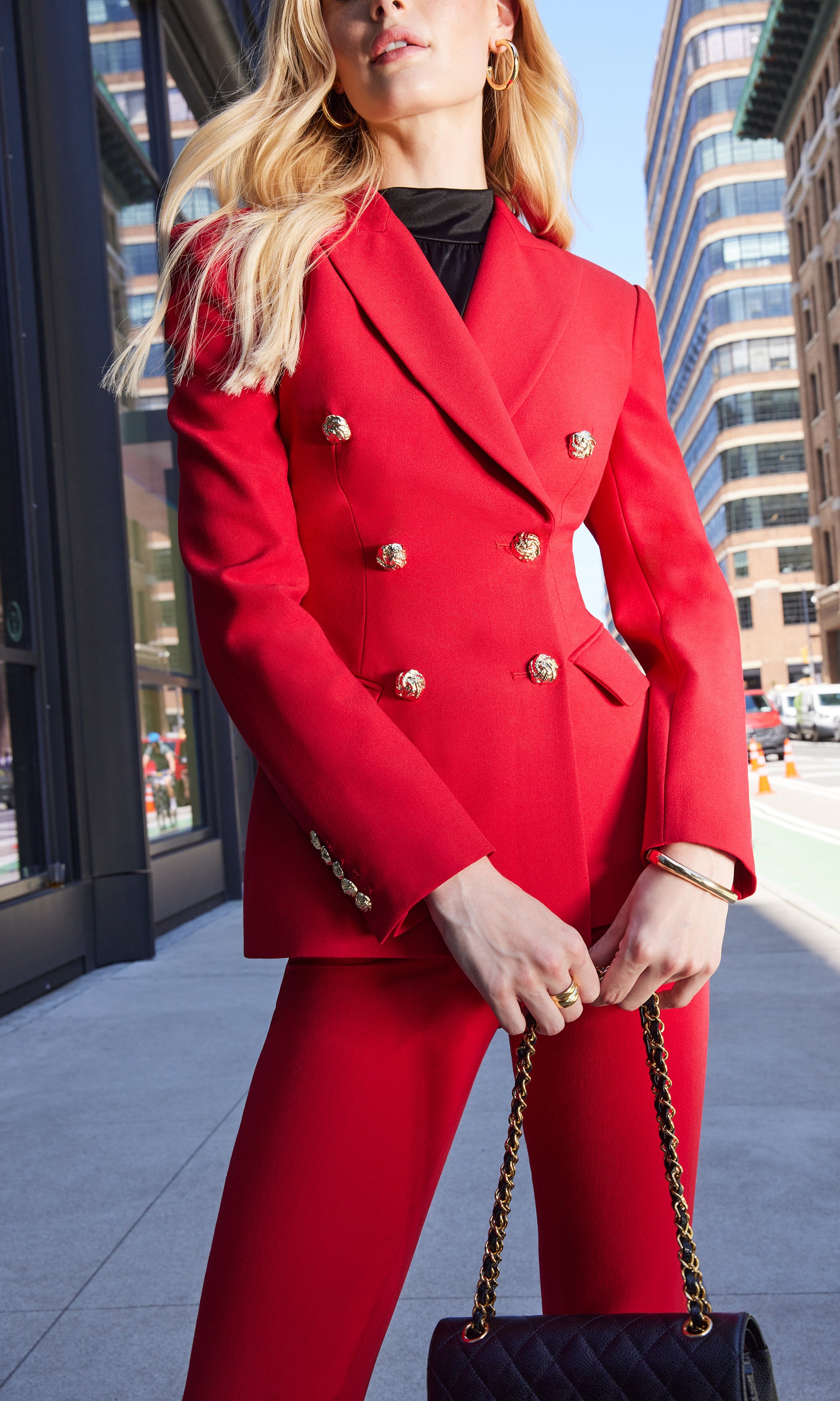 Person wearing a red suit with gold buttons holding a black purse on a city street