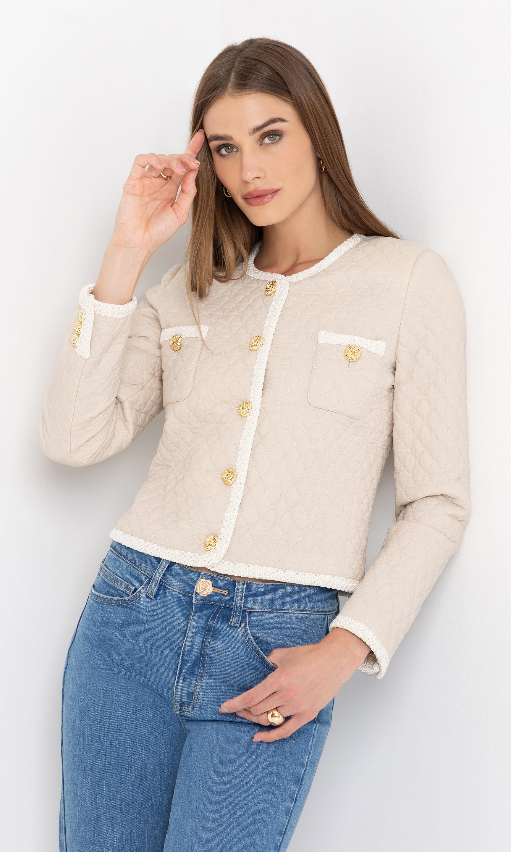 Leaning slightly with one hand near her face, she wears the cream Everie Quilted Jacket paired with high-waisted blue jeans.