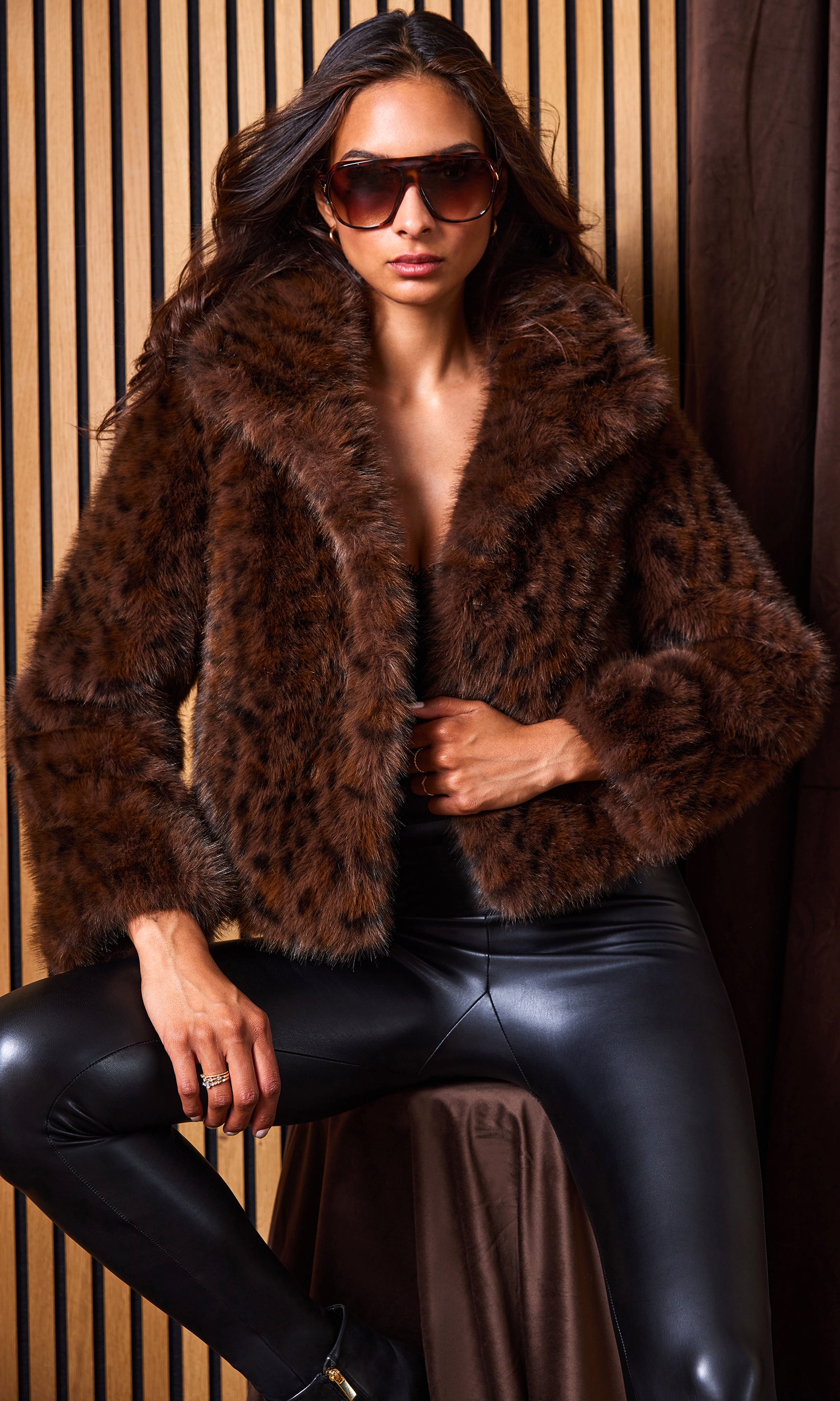 Woman wearing a brown fur coat and black leather pants against a wooden paneled wall.