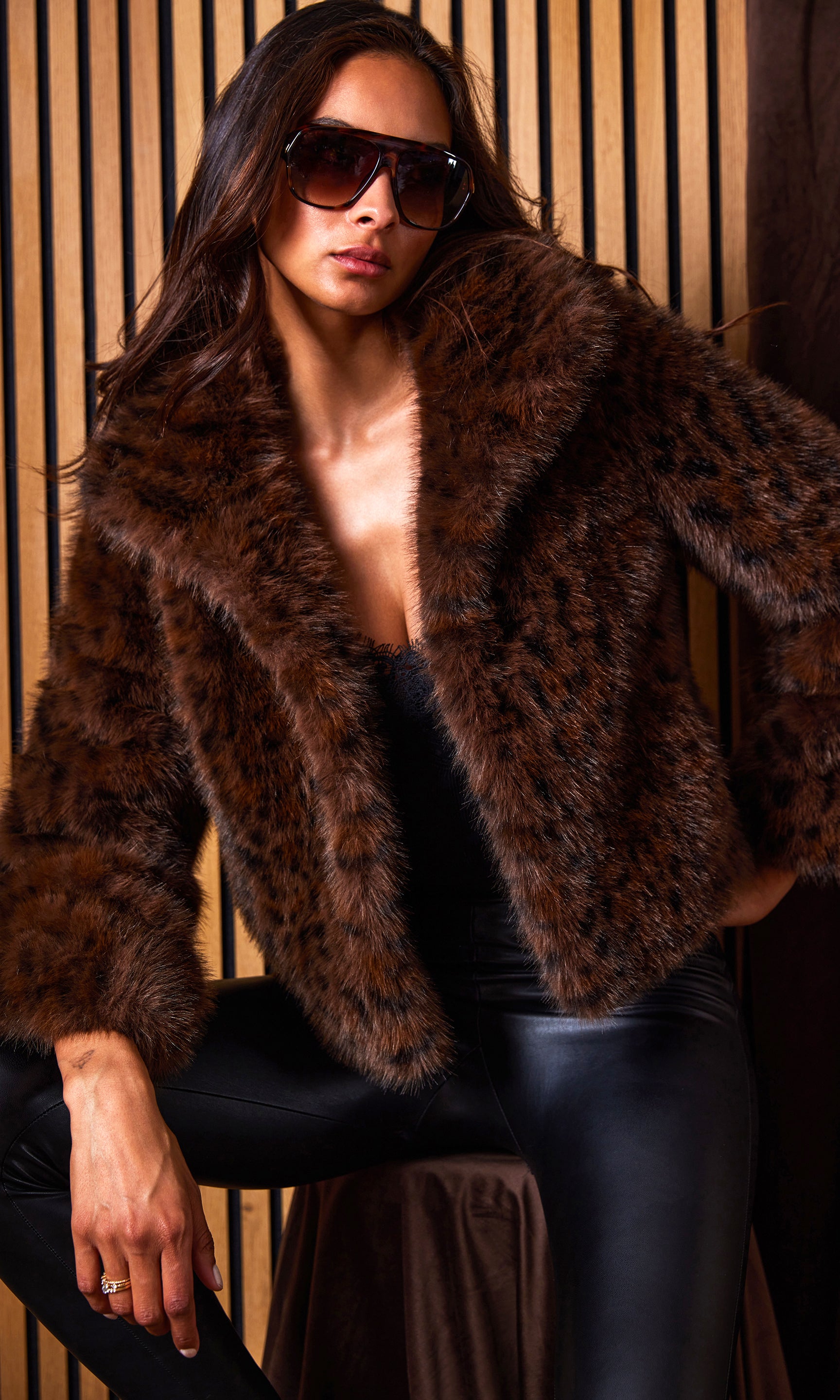 Woman wearing a brown leopard print fur coat and sunglasses against a wooden paneled wall.