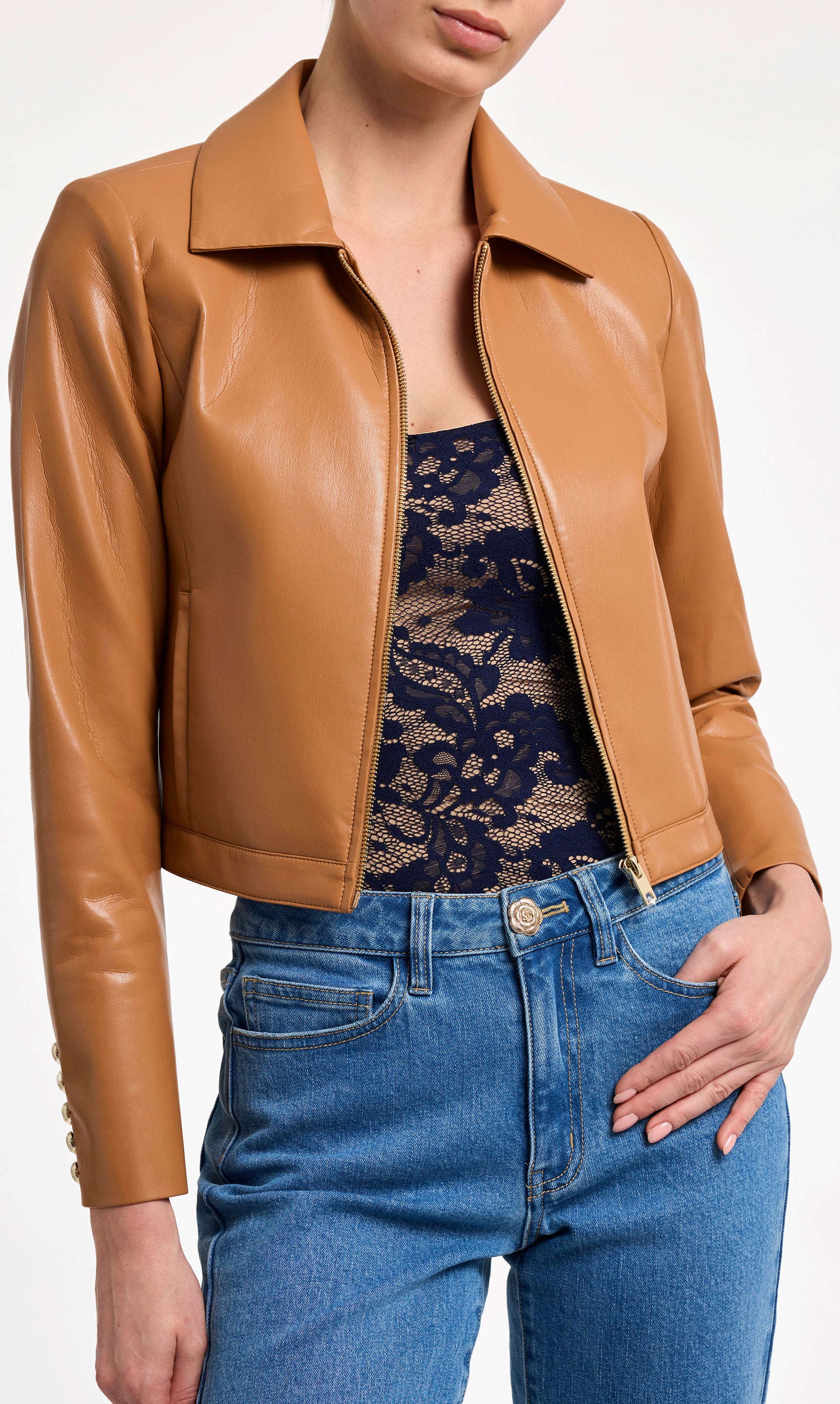 Person wearing a tan leather jacket, navy lace top, and blue jeans on a white background