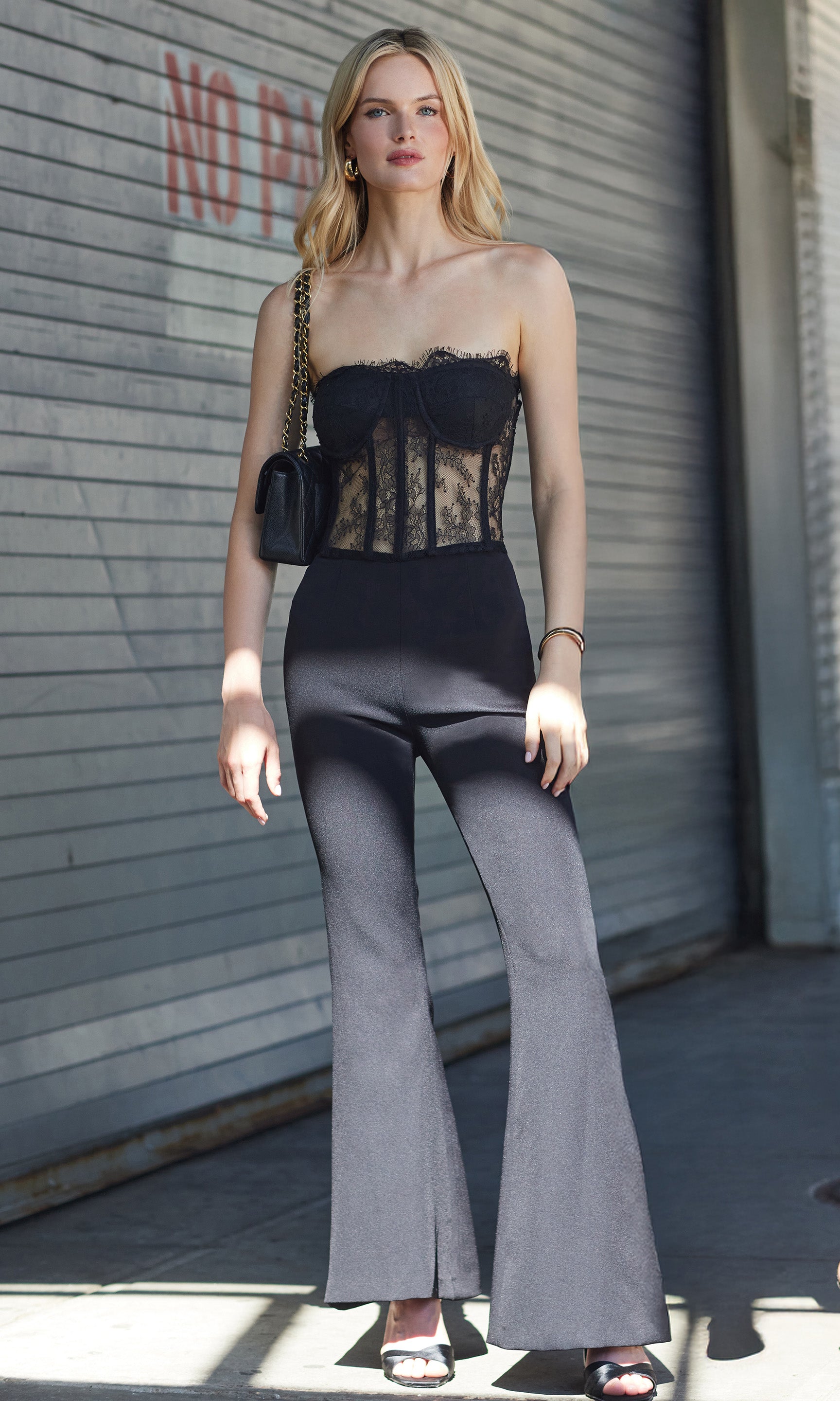 Woman wearing a black jumpsuit with a lace top and black flared pants standing in an alley.