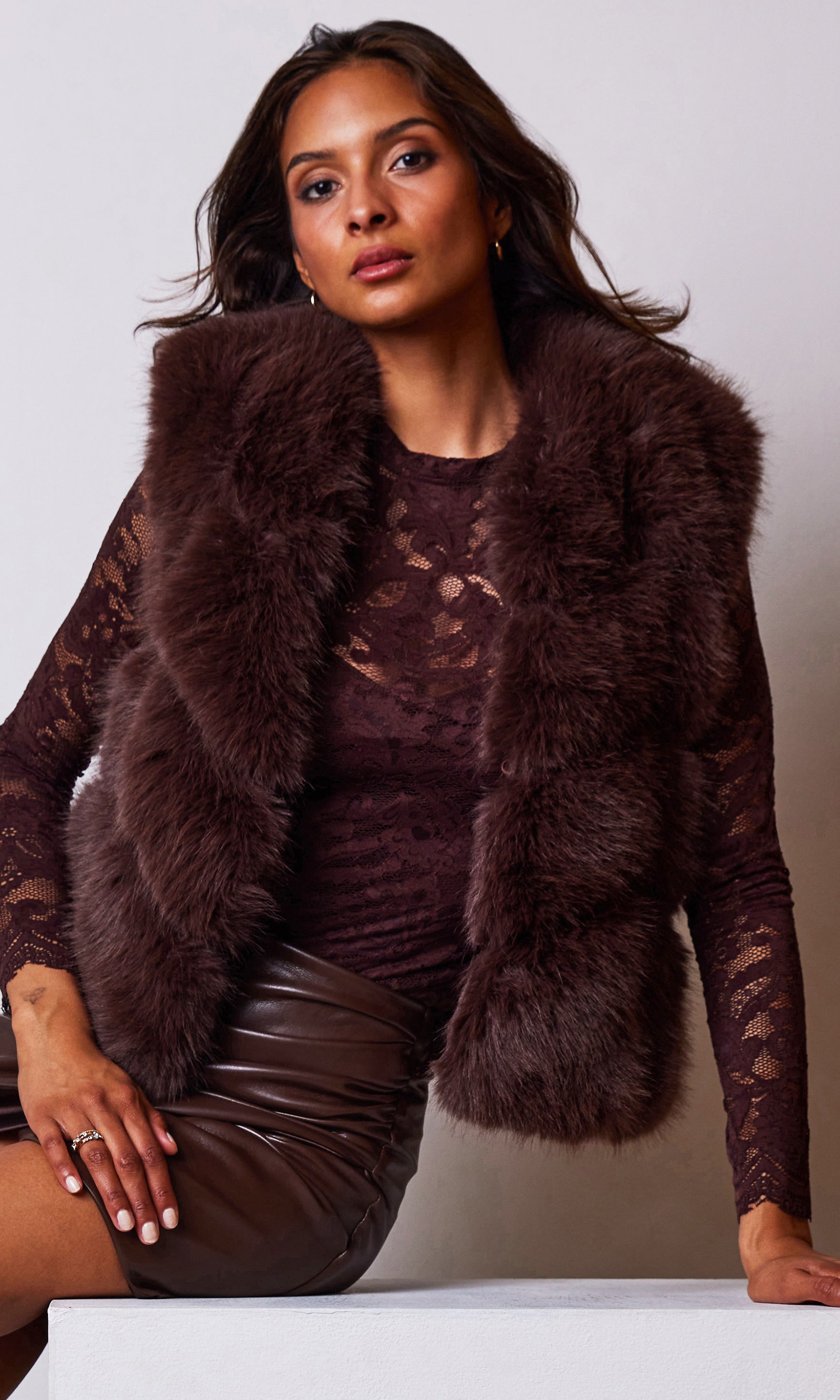 Woman wearing a brown fur vest over a lace top and skirt against a plain background