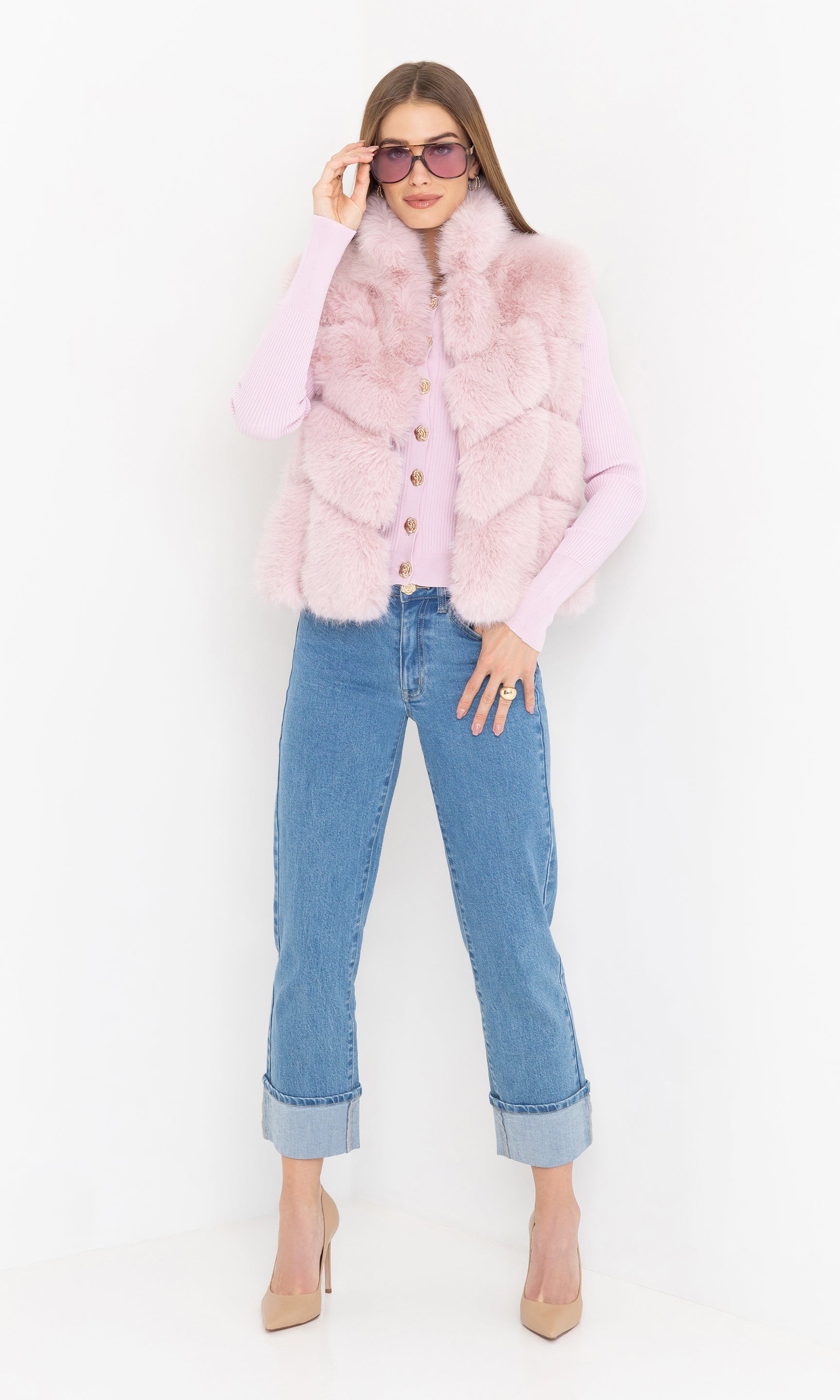 Woman wearing a pink faux fur vest, light pink shirt, blue jeans, and sunglasses on a white background
