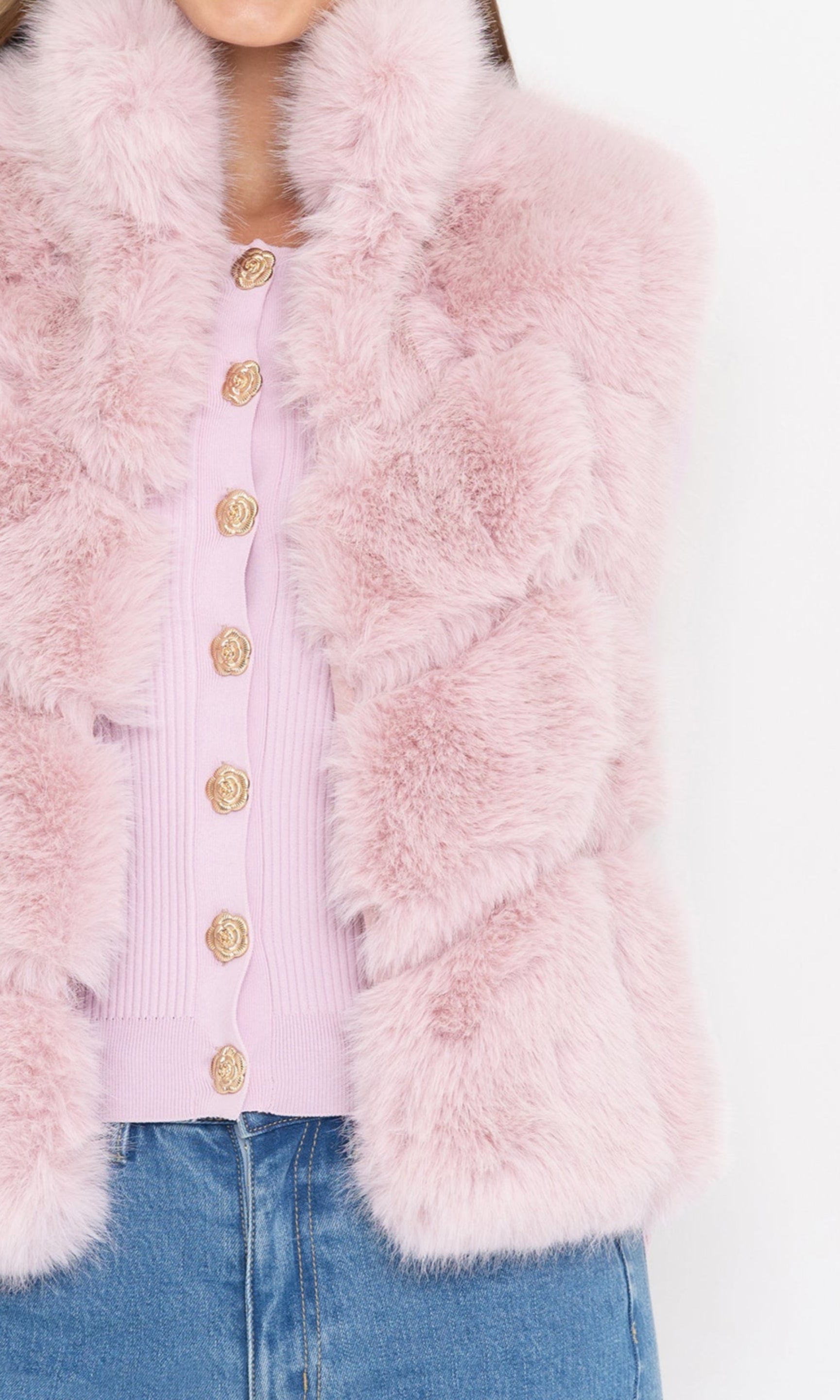 Close-up view of the model wearing the rosario faux fur vest in soft pink, paired with a light pink ribbed cardigan featuring ornate gold buttons, styled with blue denim jeans.