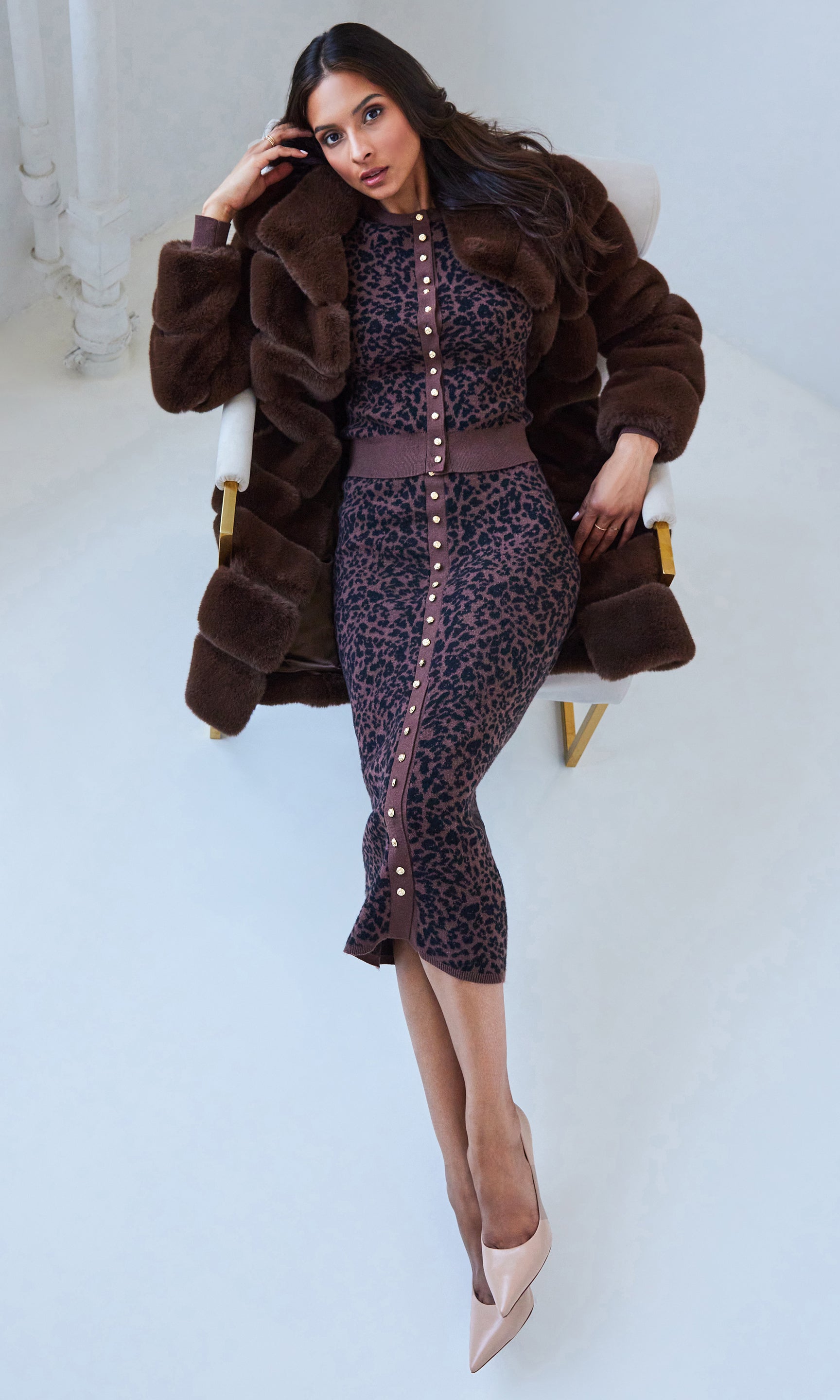 Woman wearing a leopard print outfit and brown fur coat sitting on a white chair.