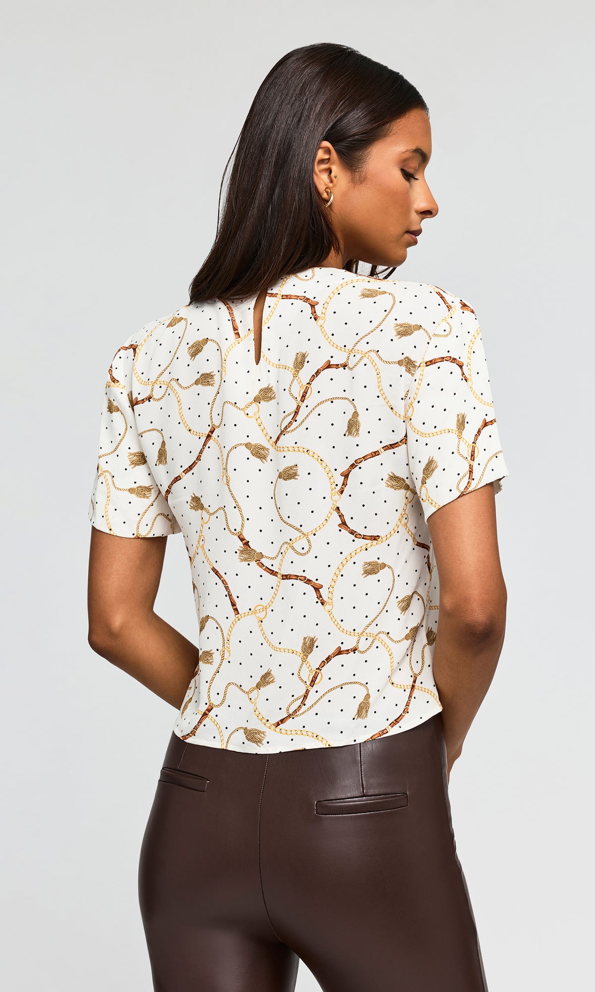 Back view of model wearing a short-sleeve top with a white base, brown and gold tassel pattern.