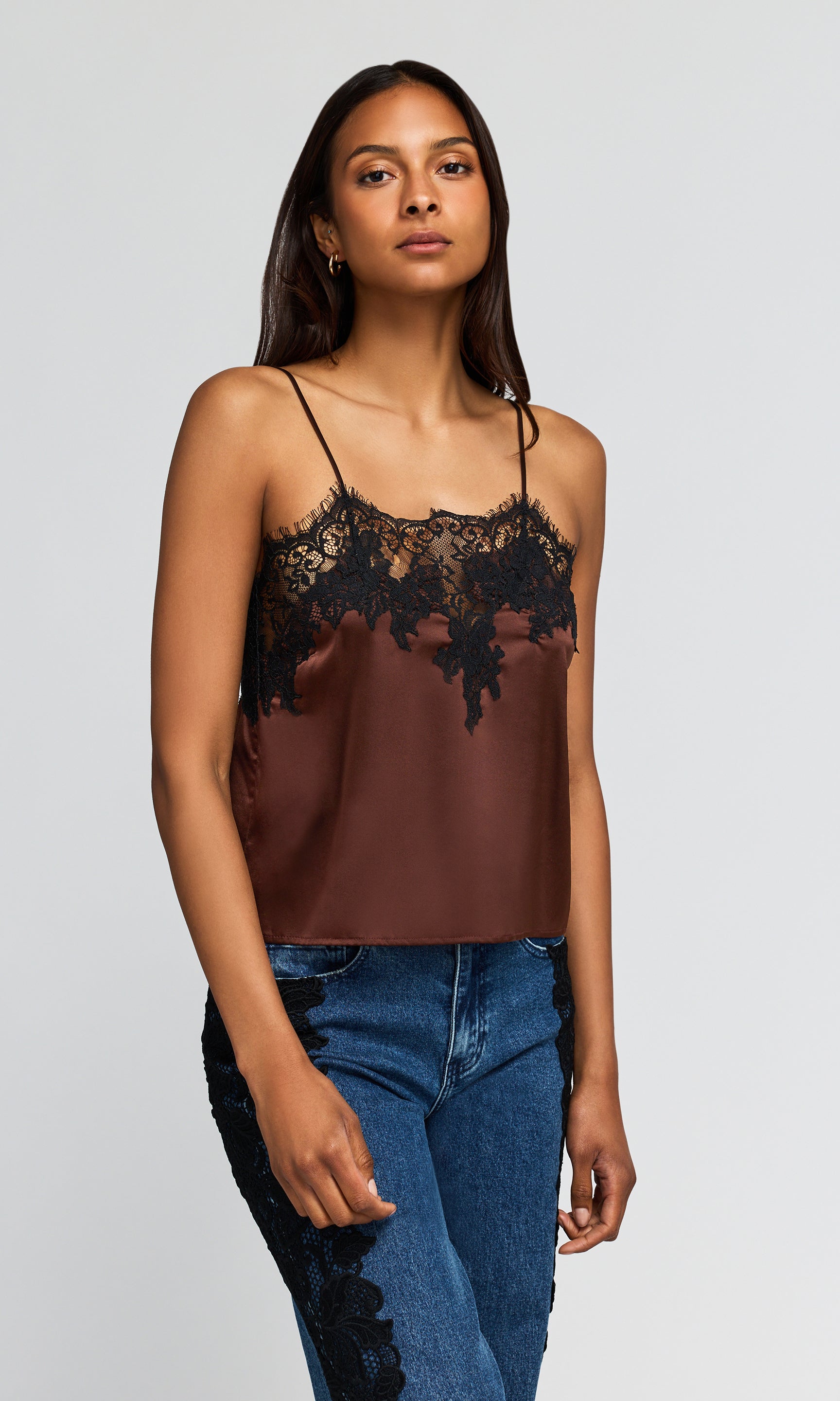 Side view of model wearing a brown satin camisole top with black lace trim along the neckline, paired with blue jeans.