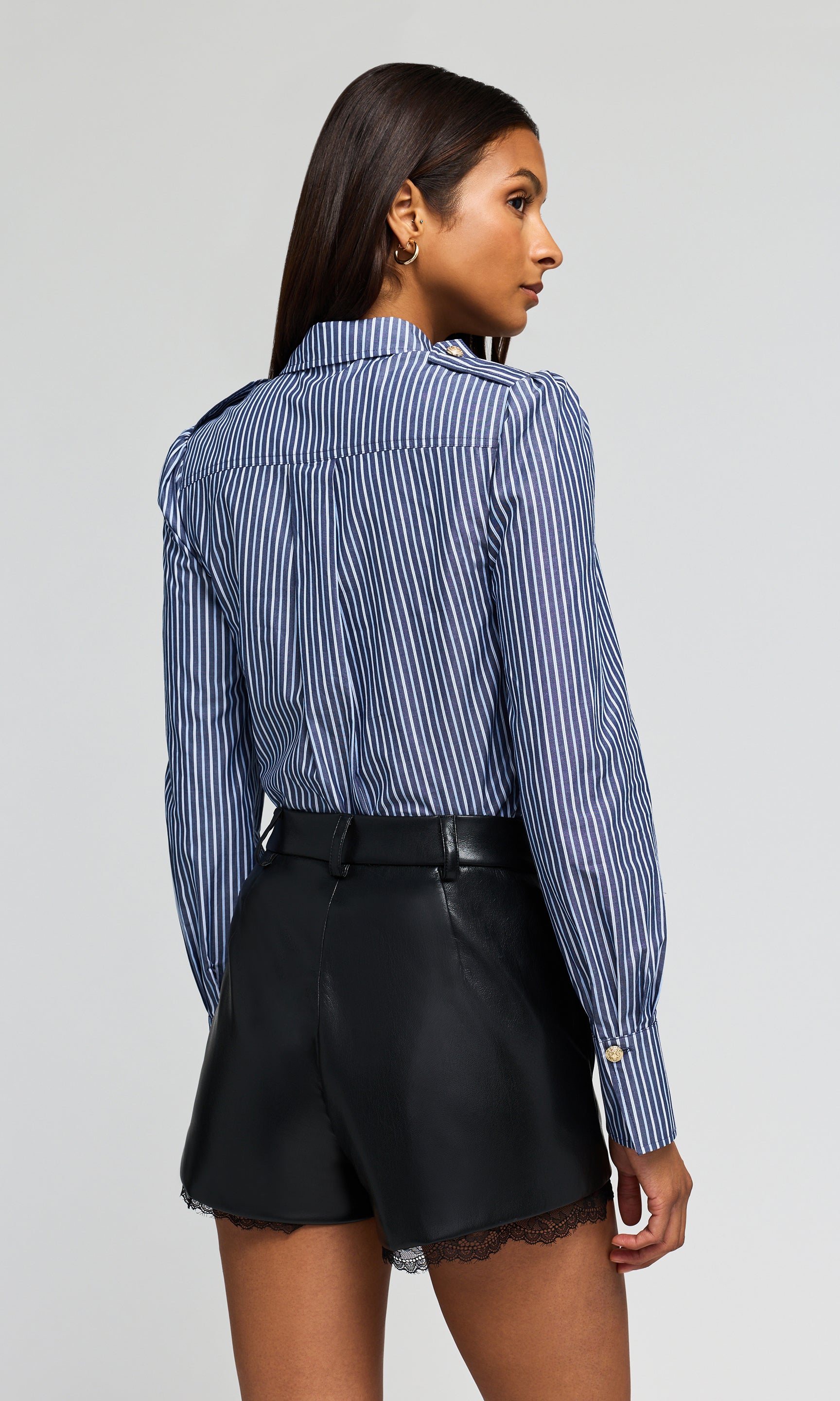 Back view of model wearing a blue and white striped button-up shirt tucked into black shorts, on a plain background.