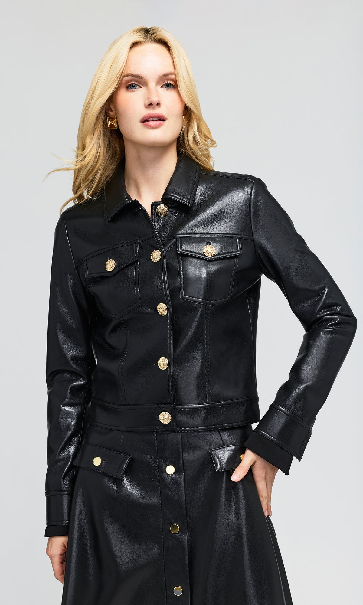 Model wearing a black faux-leather jacket and matching skirt set, featuring gold-tone buttons and flap pockets.