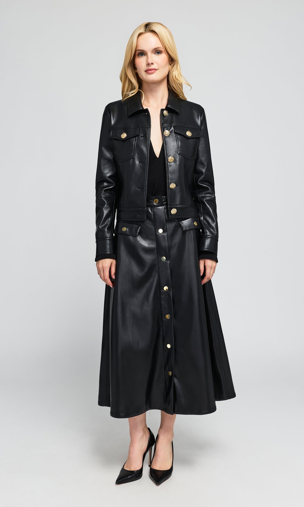 Model wearing a black faux-leather longline jacket and matching midi skirt set with gold-tone buttons.