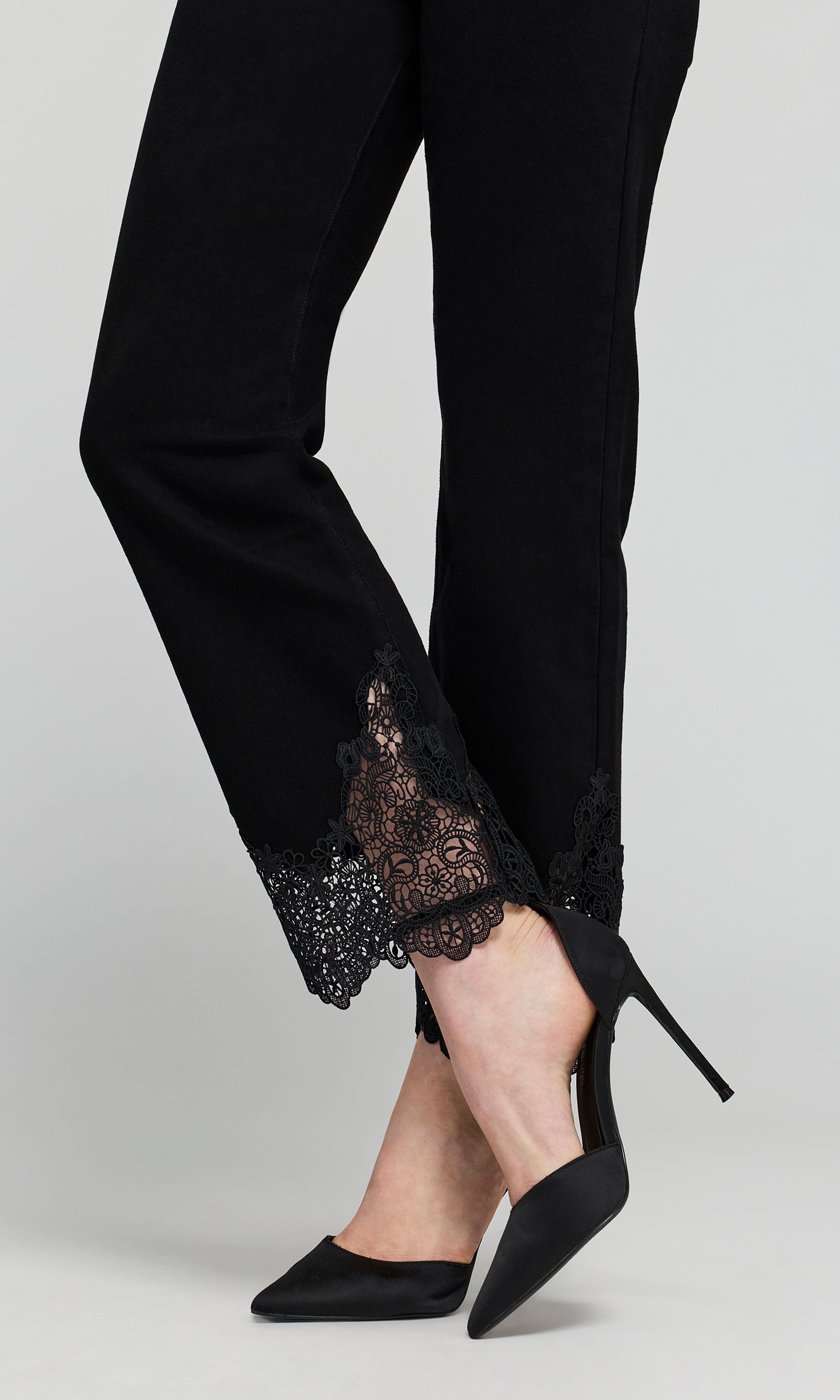 Close-up of black jeans with a sheer black lace trim at the hem, shown on a model on a plain background.