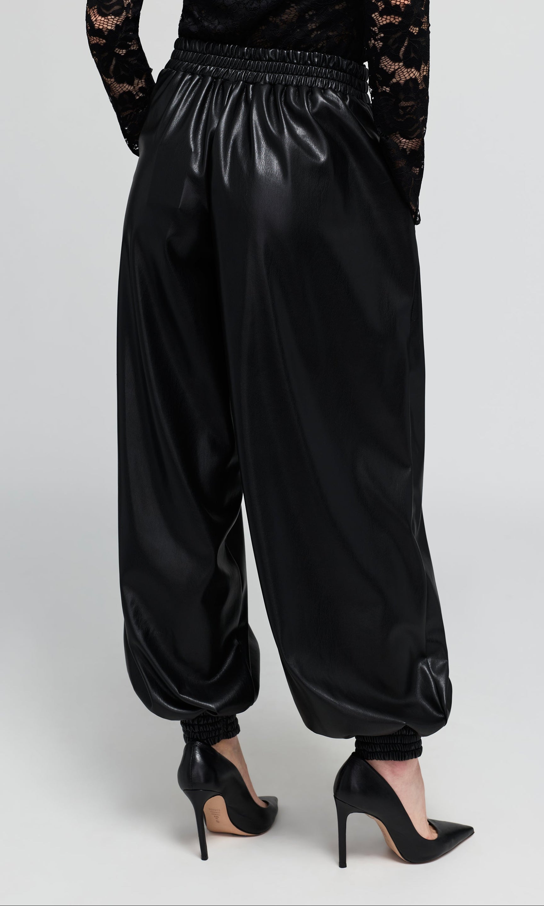 Back view of black vegan-leather, wide-leg harem pants with an elastic waistband, and gathered cuffs, shown on a model.