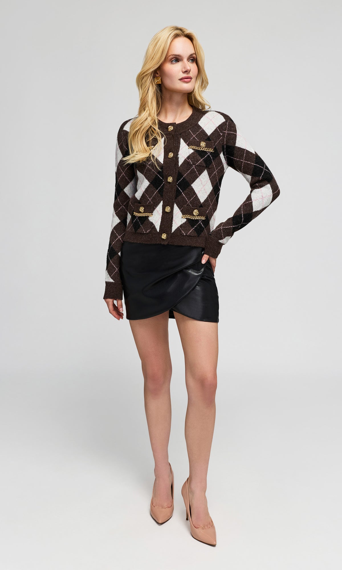 Model wearing a brown and white argyle cardigan with gold buttons, a black faux-leather mini skirt, and beige heels.