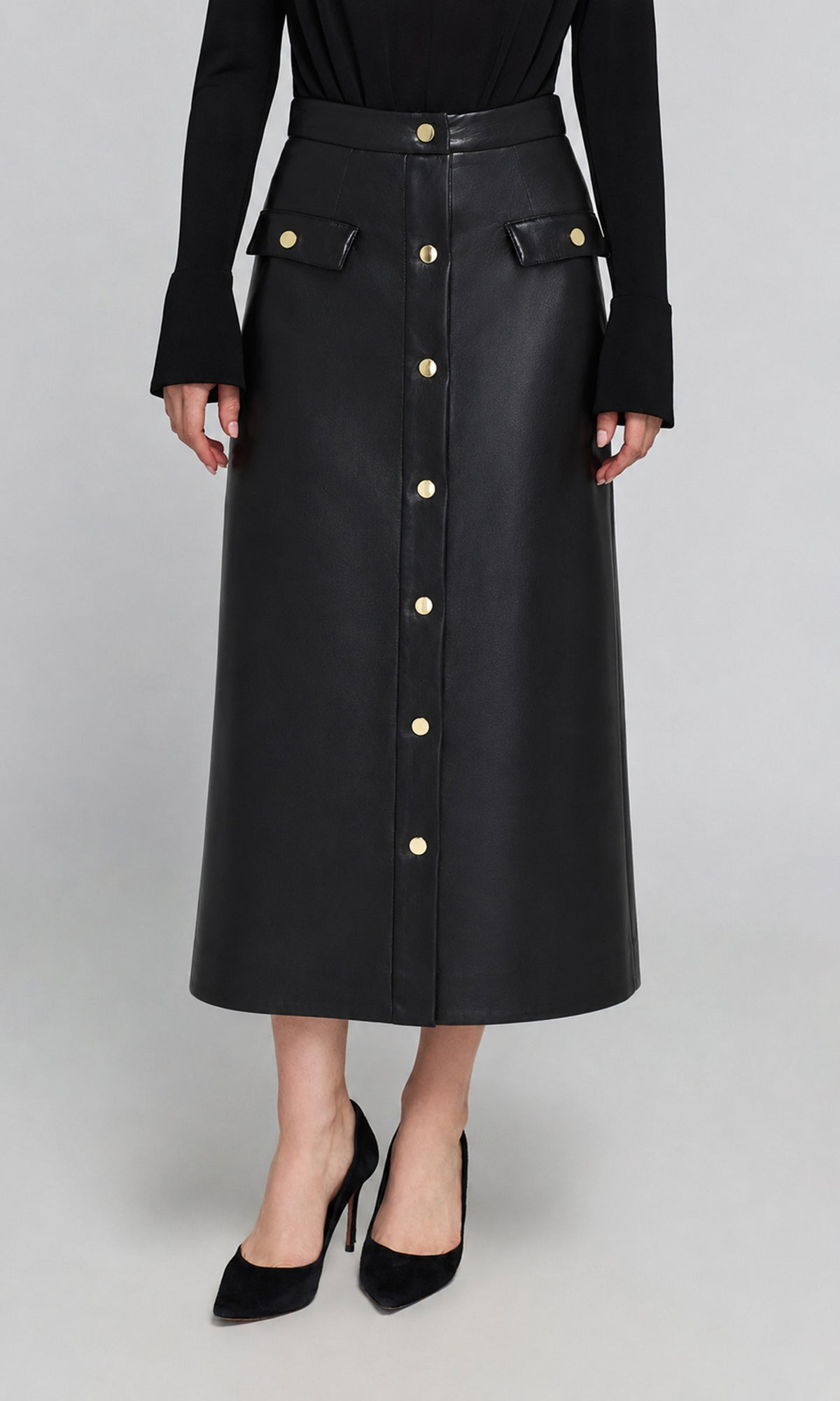 Model wearing a black faux-leather midi skirt with gold-tone button details and flap pockets, paired with a black long-sleeve top and black high heels.