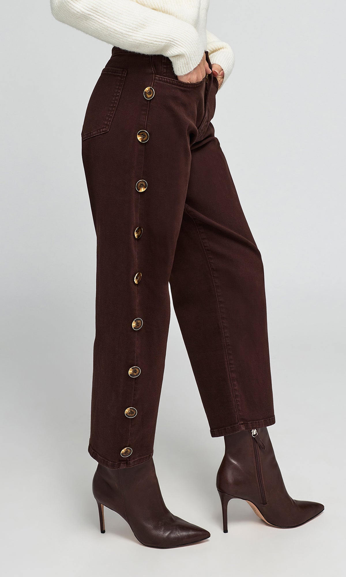 Side view of model wearing dark brown cropped jeans with decorative gold-tone side buttons, paired with a cream sweater.