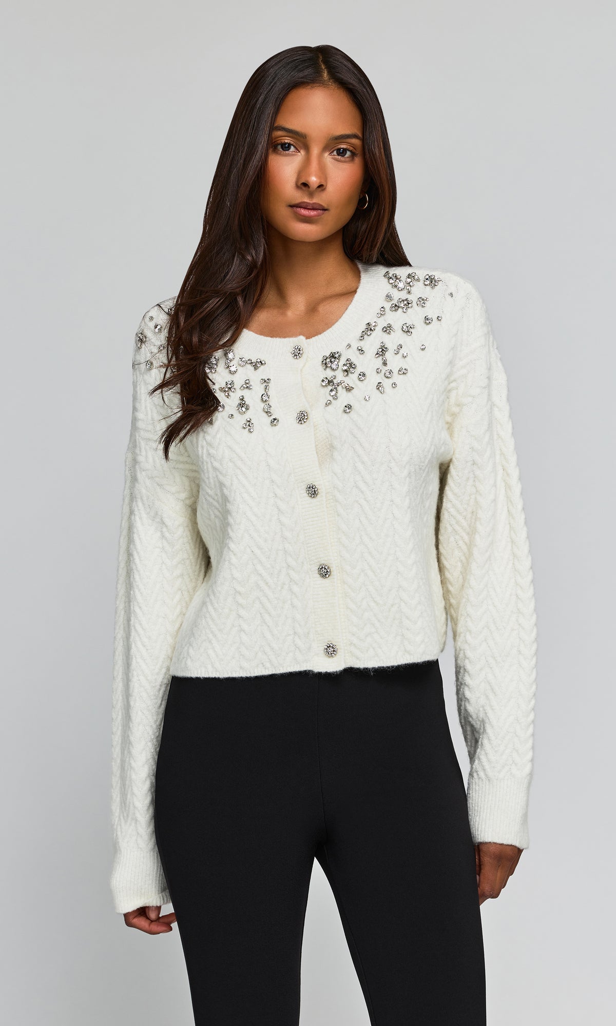 Model wearing a cream cable-knit cropped cardigan with silver floral embroidery and button closures, paired with black pants.