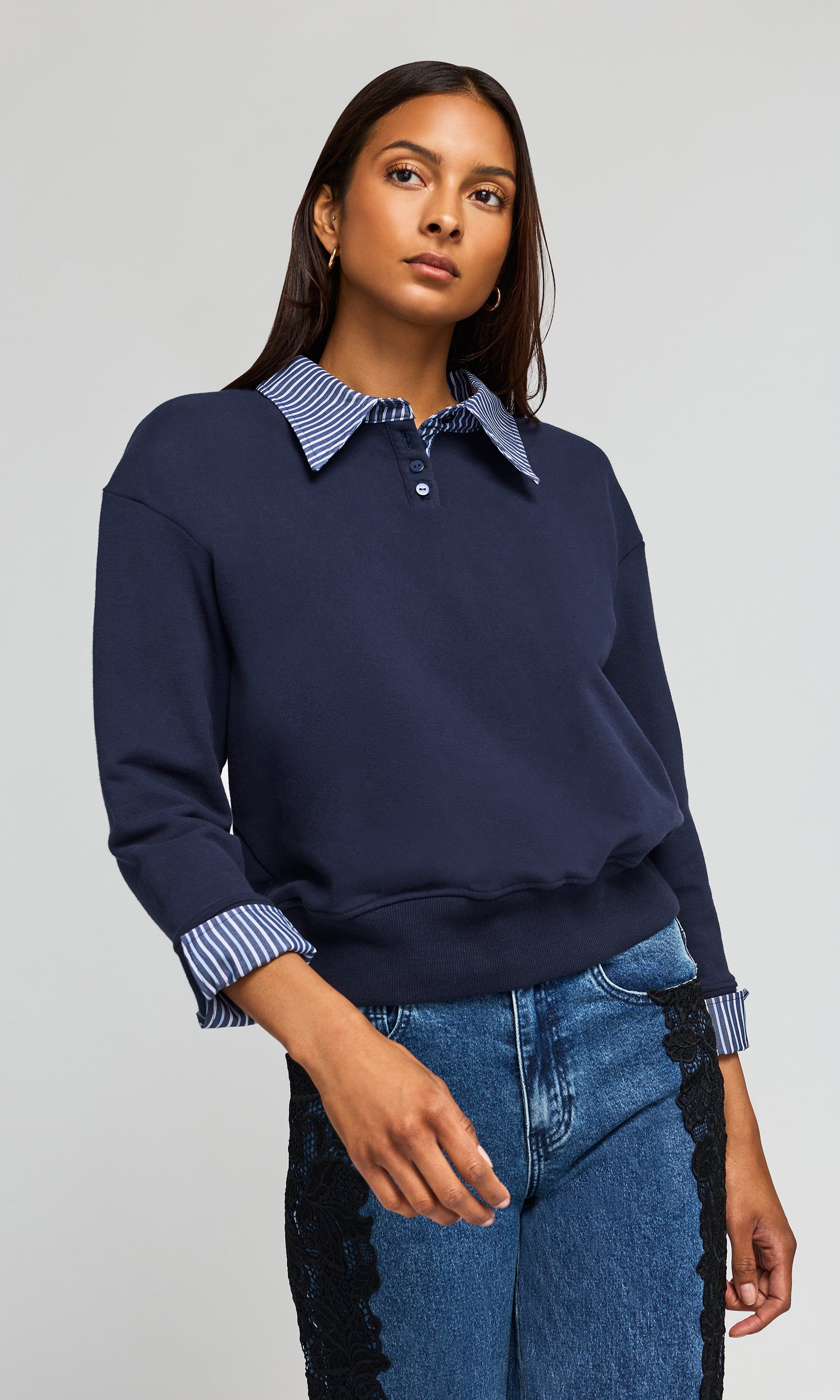 Side view of model wearing a navy blue long-sleeve sweatshirt with a blue and white striped polo collar, paired with blue jeans, against a light gray background.