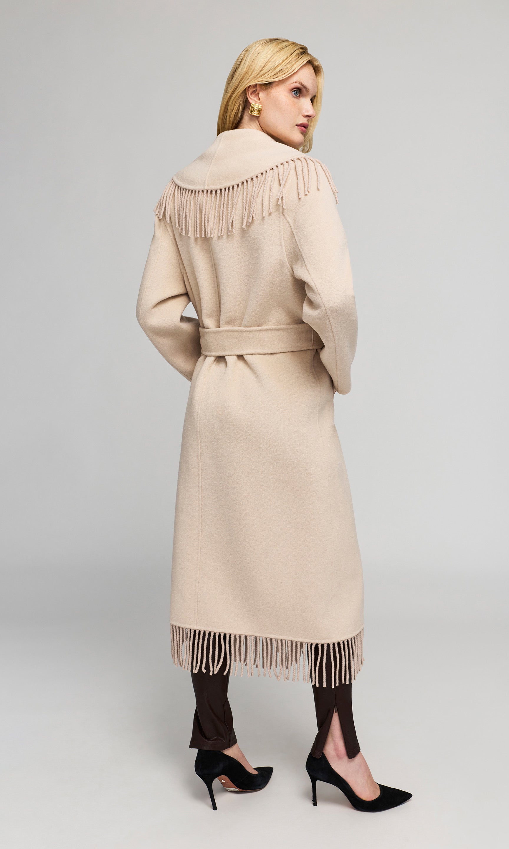 Back view of model wearing a long beige coat with fringe detailing on the shoulders and hem, paired with dark brown tights and black heels.