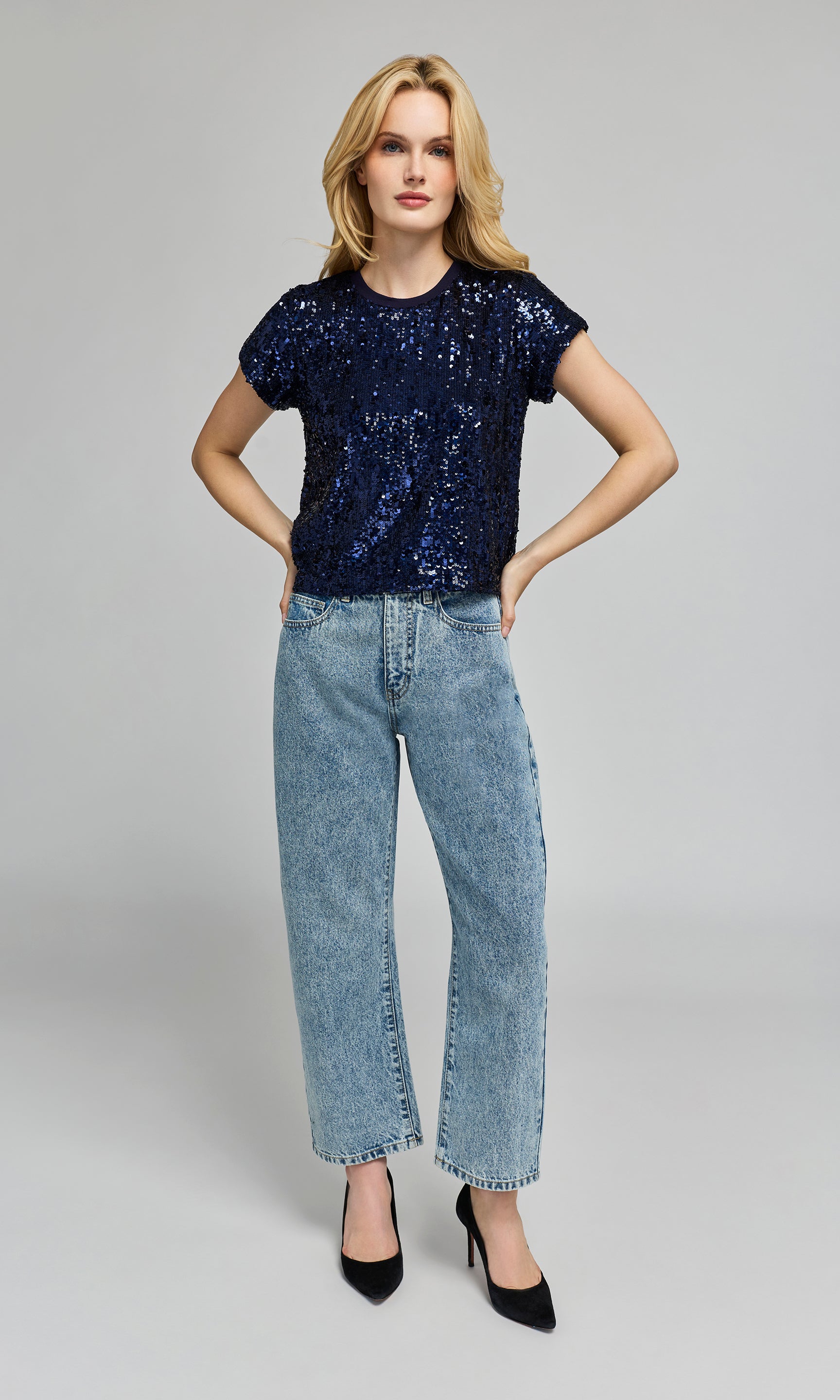 Woman wearing a navy sequin top and light blue jeans on a gray background