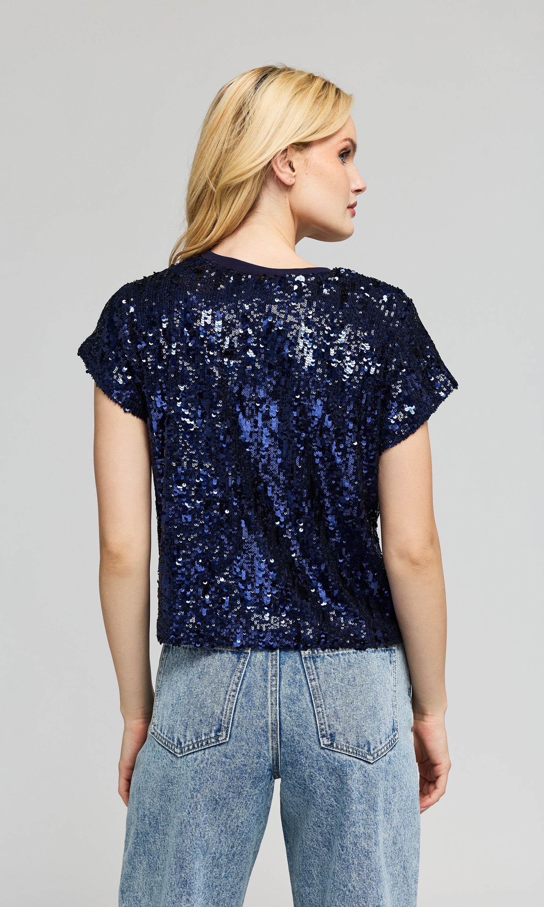 Back view of woman wearing a navy sequin top and light blue jeans on a gray background