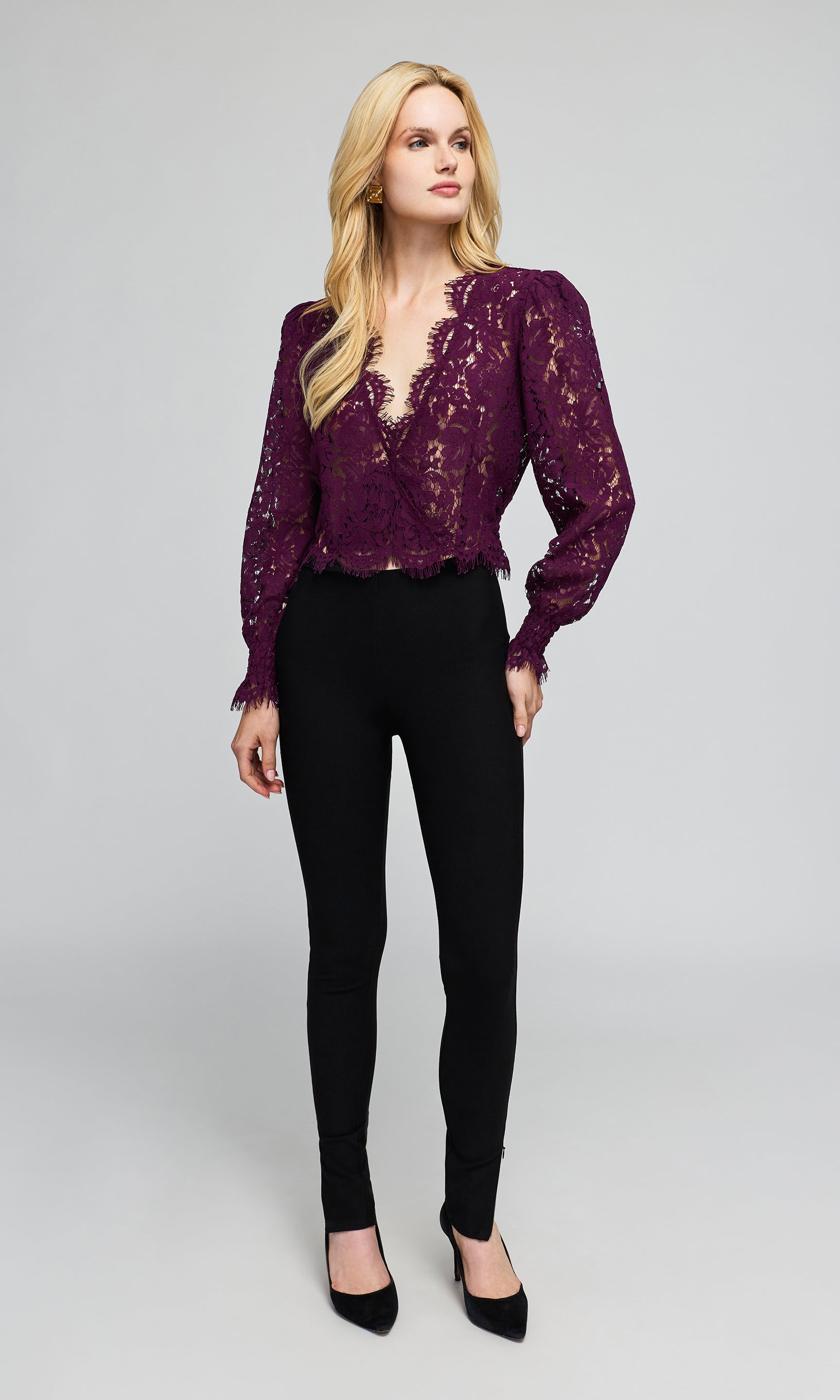 A blonde model wearing a deep purple lace long-sleeve blouse with puff sleeves and black skinny pants, on a plain background.