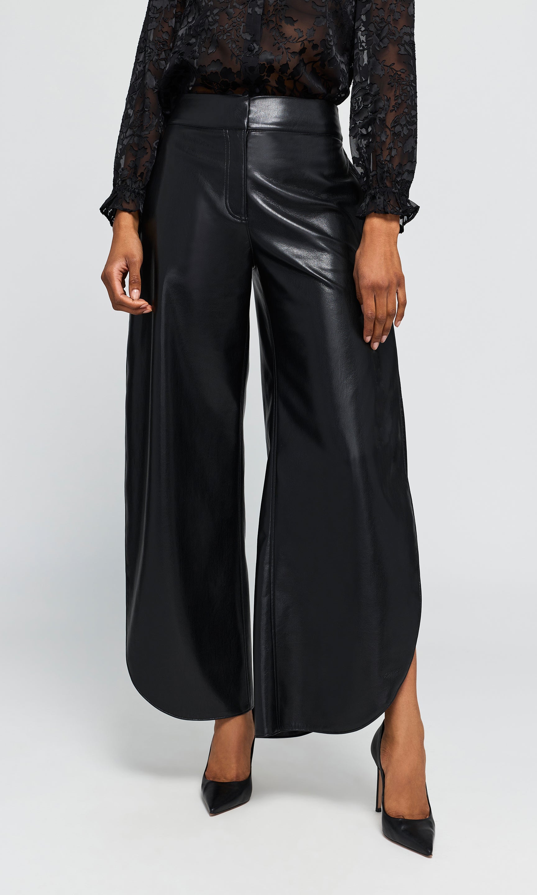 Model wearing black wide-leg trousers with a glossy faux-leather finish, paired with a black lace long-sleeve top and black pointed-toe heels.