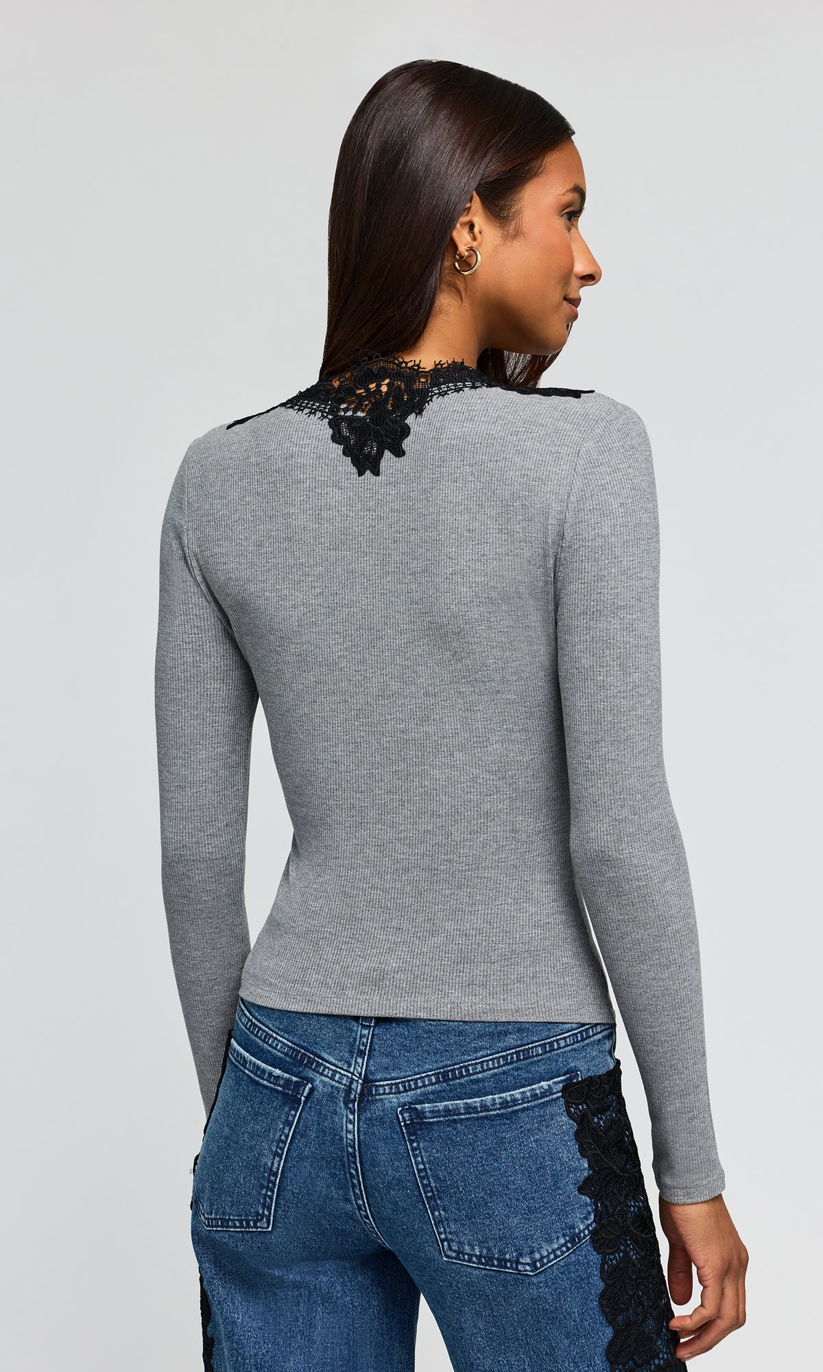 Back view of model wearing a fitted long-sleeve gray top with black lace trim around the scoop neckline, paired with blue jeans featuring black lace side panels.