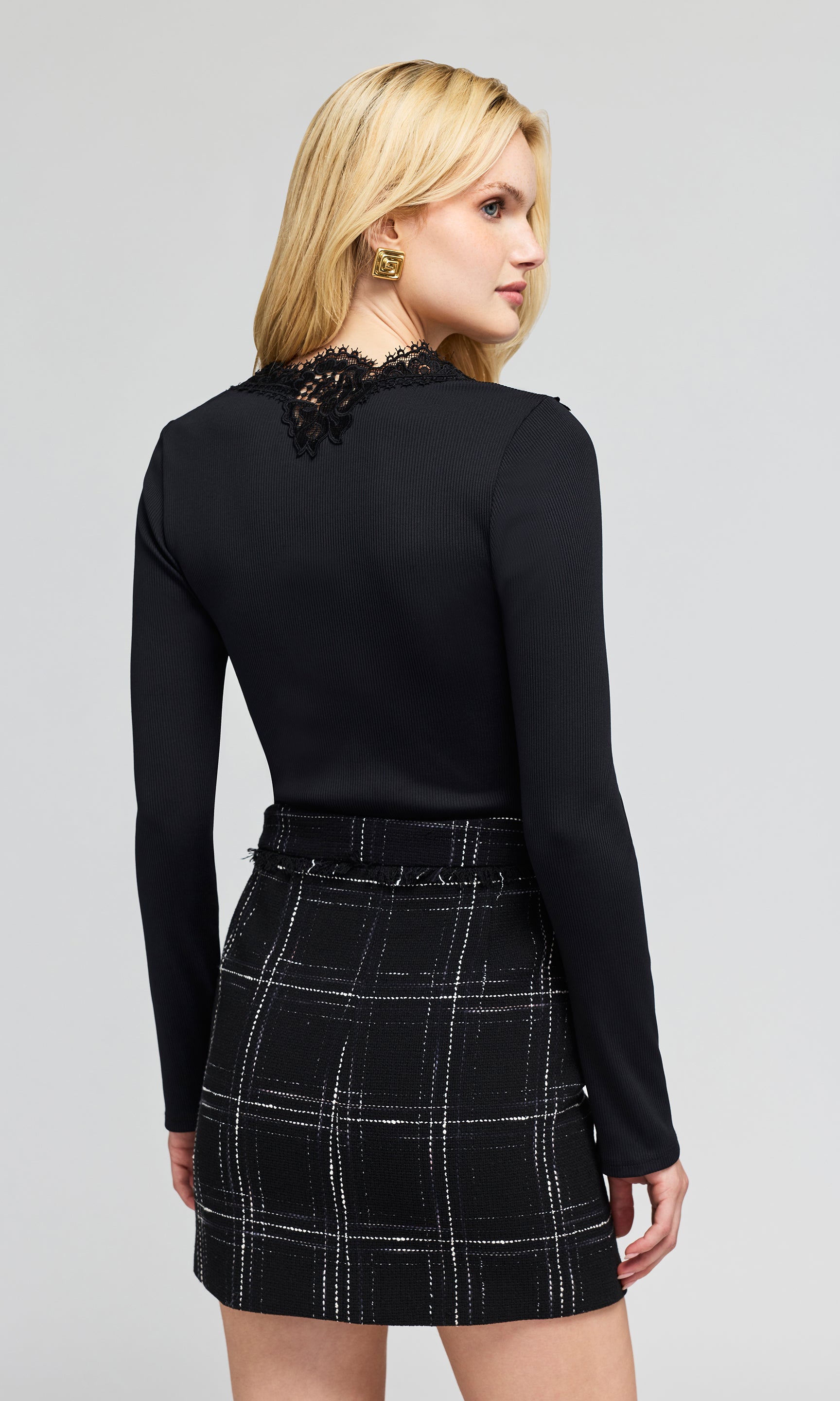 Back view of model wearing a black long-sleeve knit top with a lace V-neckline detail and a black plaid mini skirt.