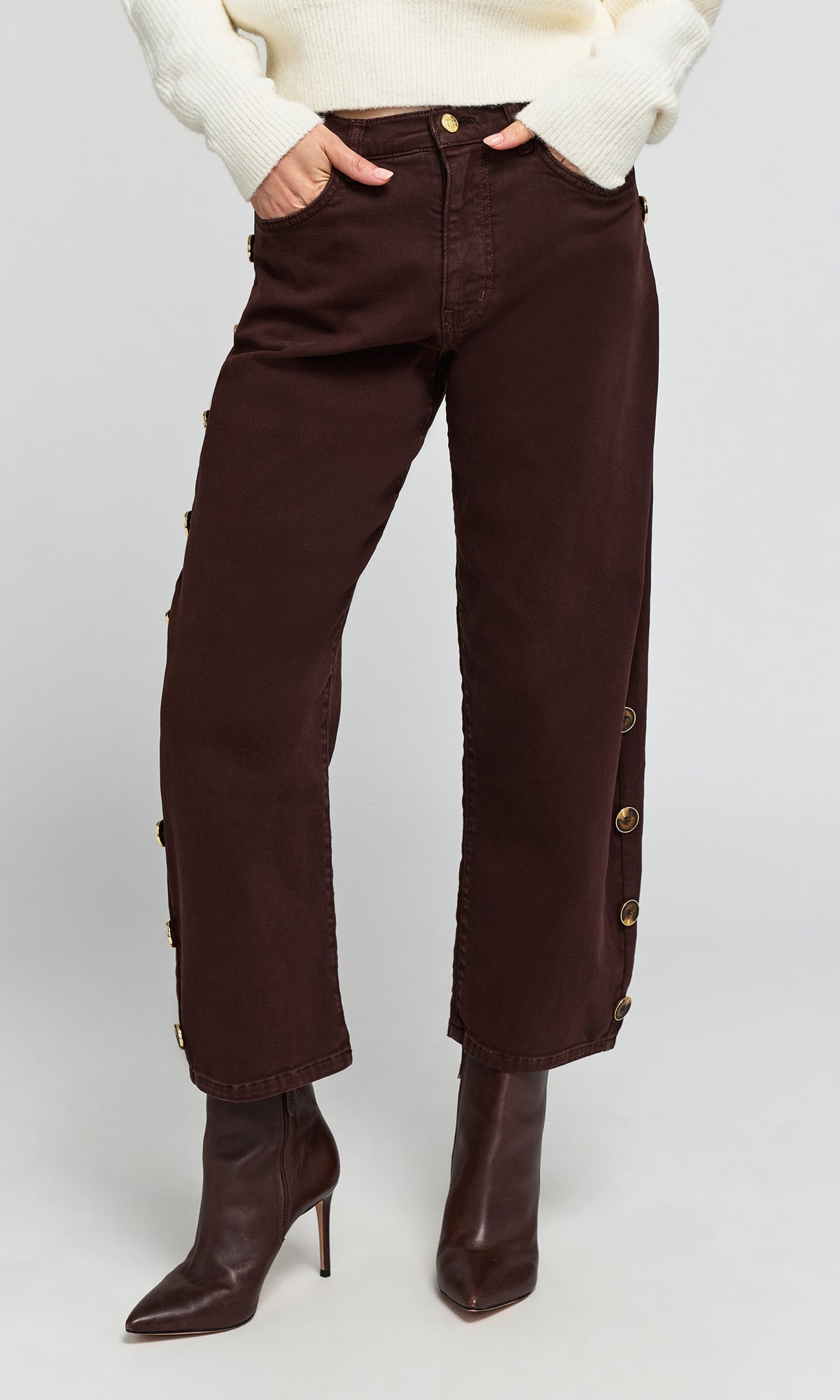 Model wearing dark brown cropped jeans with decorative gold-tone side buttons, paired with a cream sweater.