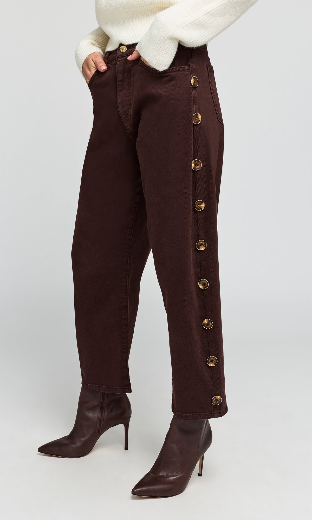 Side view of model wearing dark brown cropped jeans with decorative gold-tone side buttons, paired with a cream sweater.
