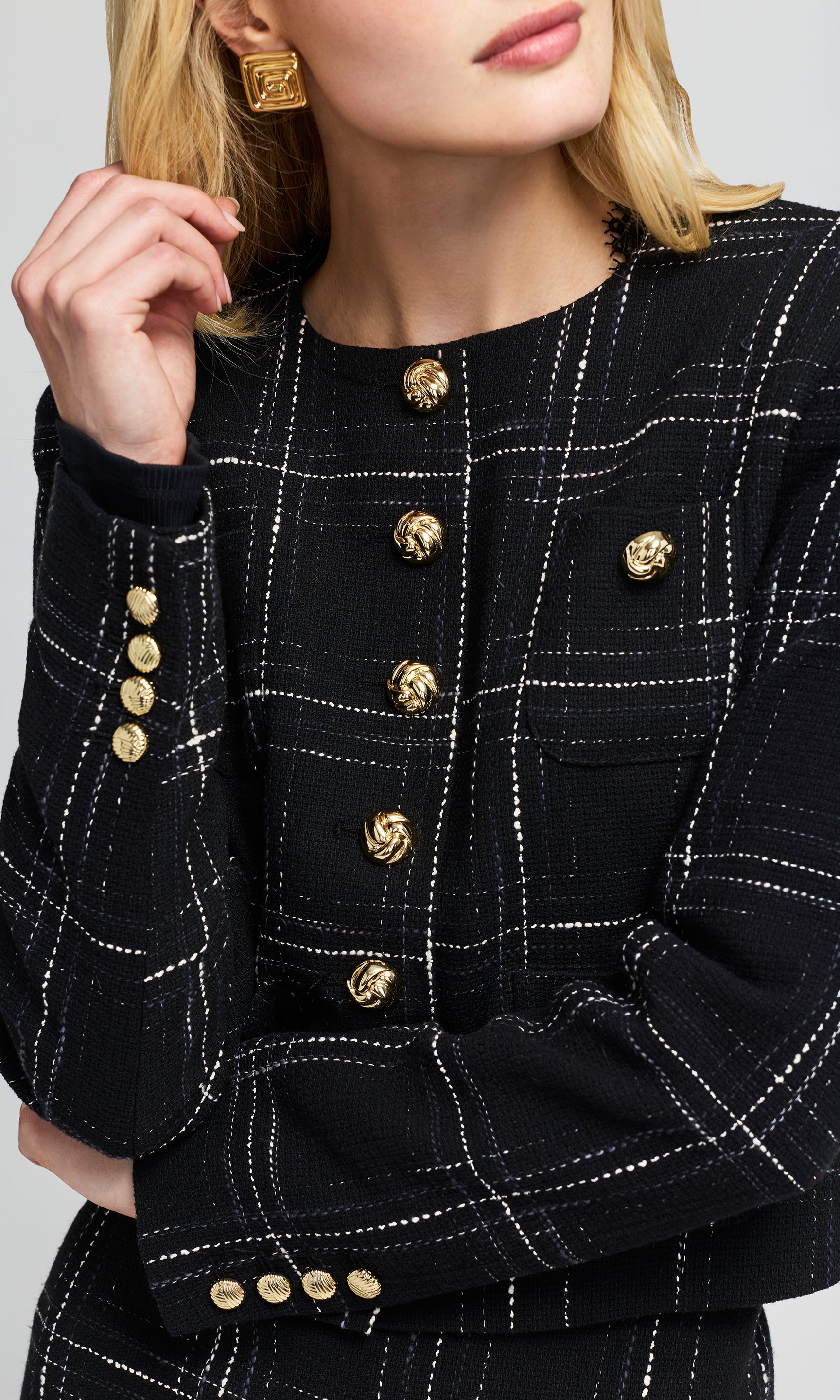Close-up of a model wearing a black tweed blazer with gold buttons and white windowpane plaid lines.
