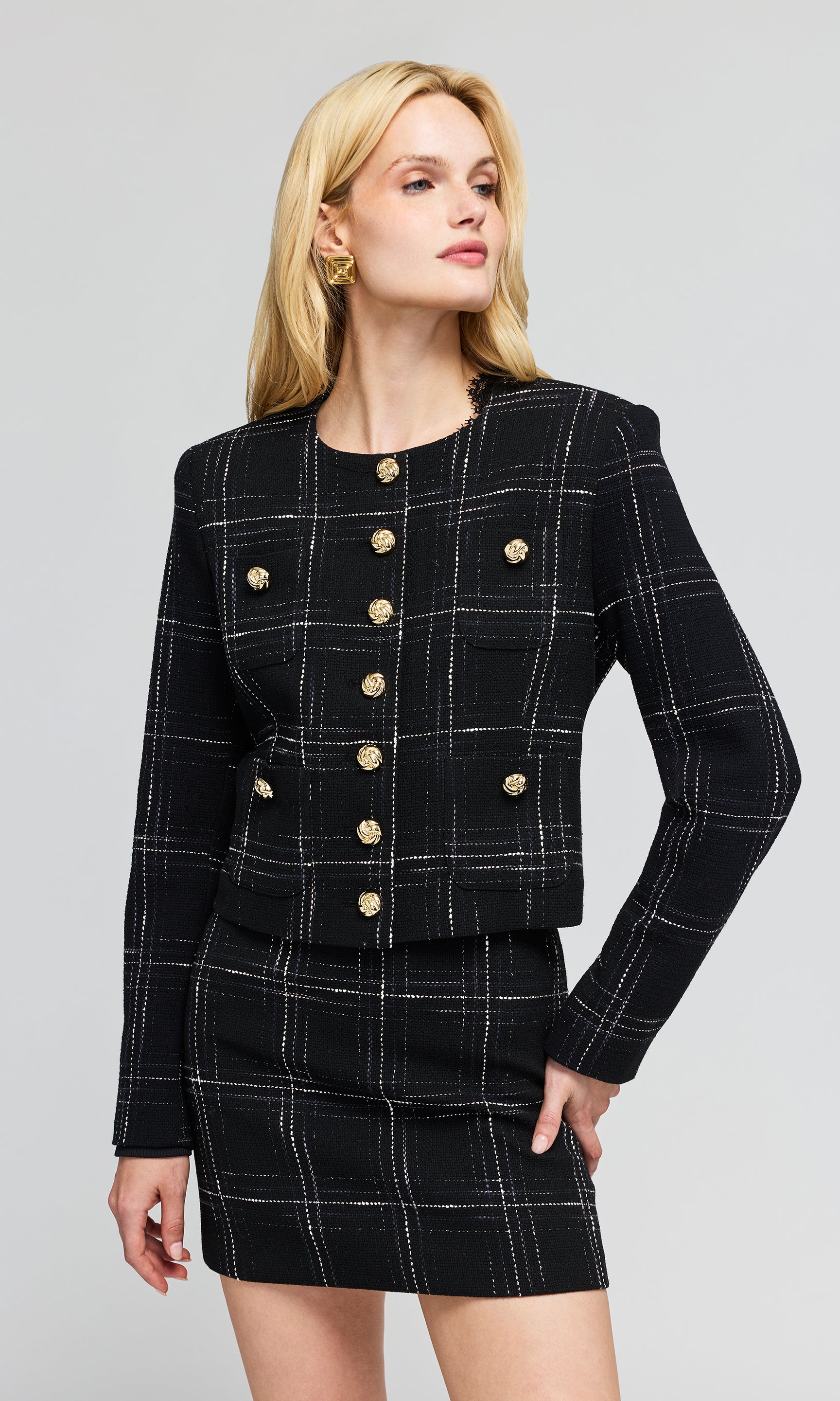A blonde model wearing a black and white plaid tweed jacket and matching mini skirt with gold buttons.