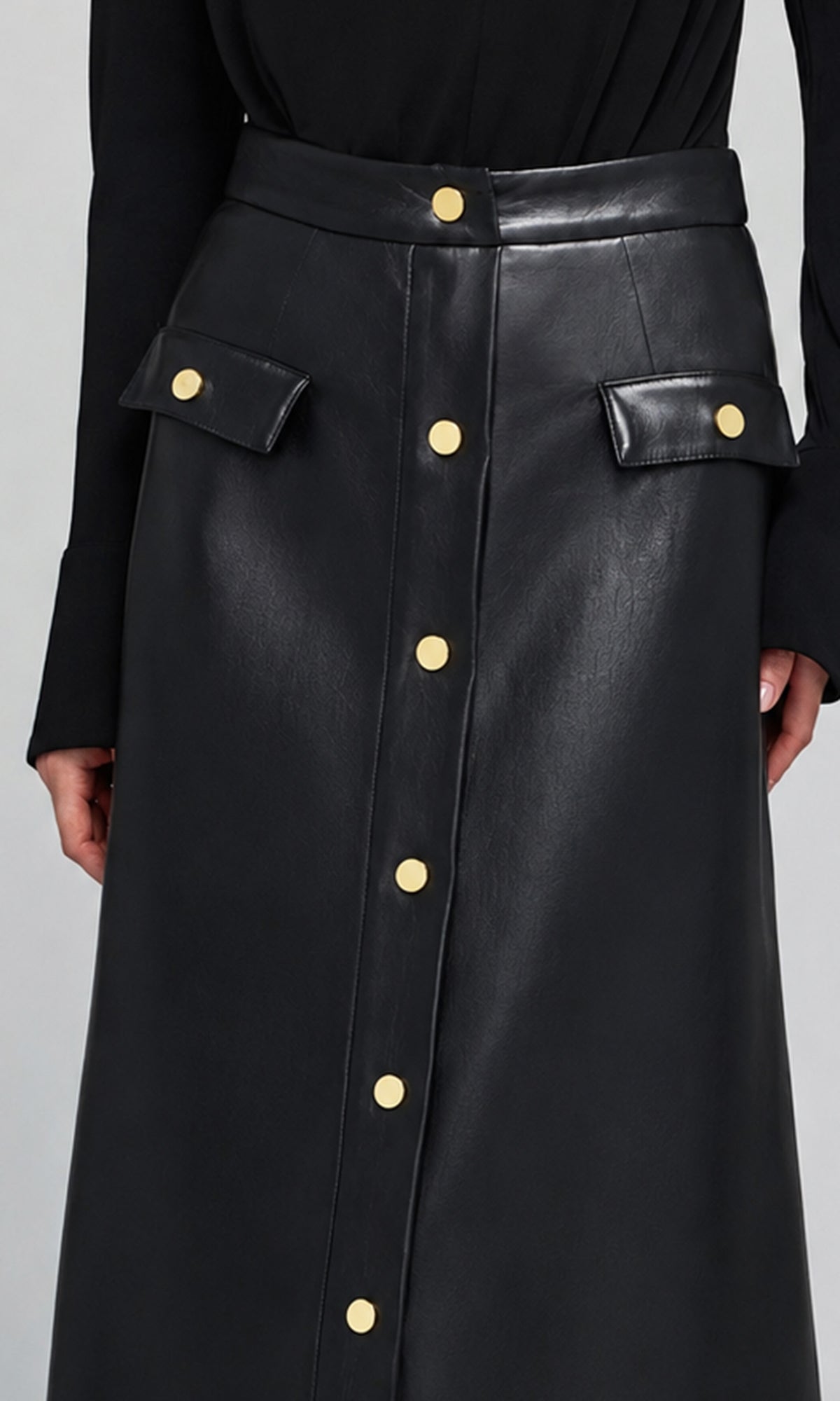 A close-up of a black faux-leather midi skirt with gold-tone button details and flap pockets.