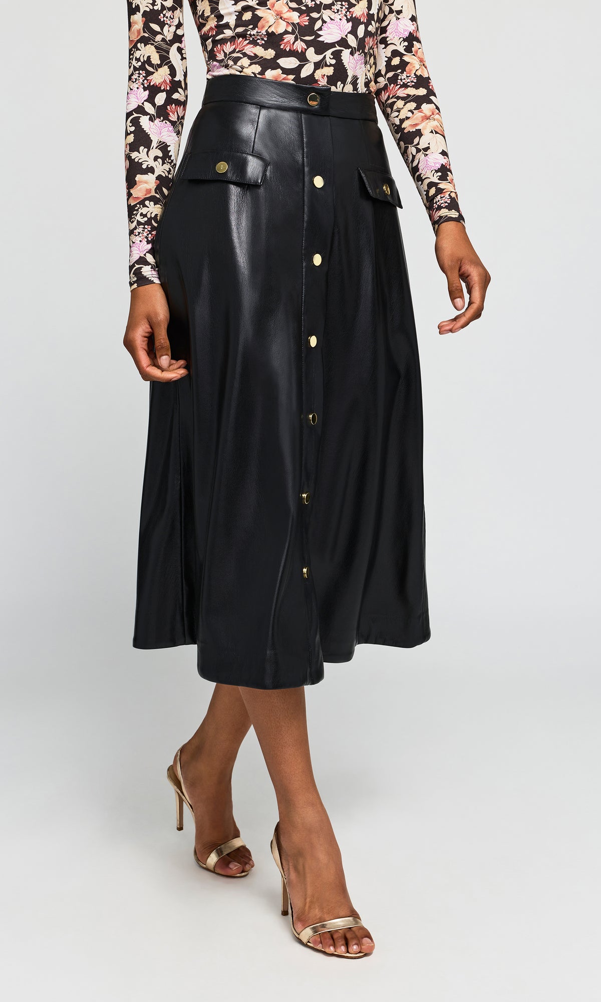 Side view of model wearing a black faux-leather midi skirt with gold button details and flap pockets, paired with a floral long-sleeve top.