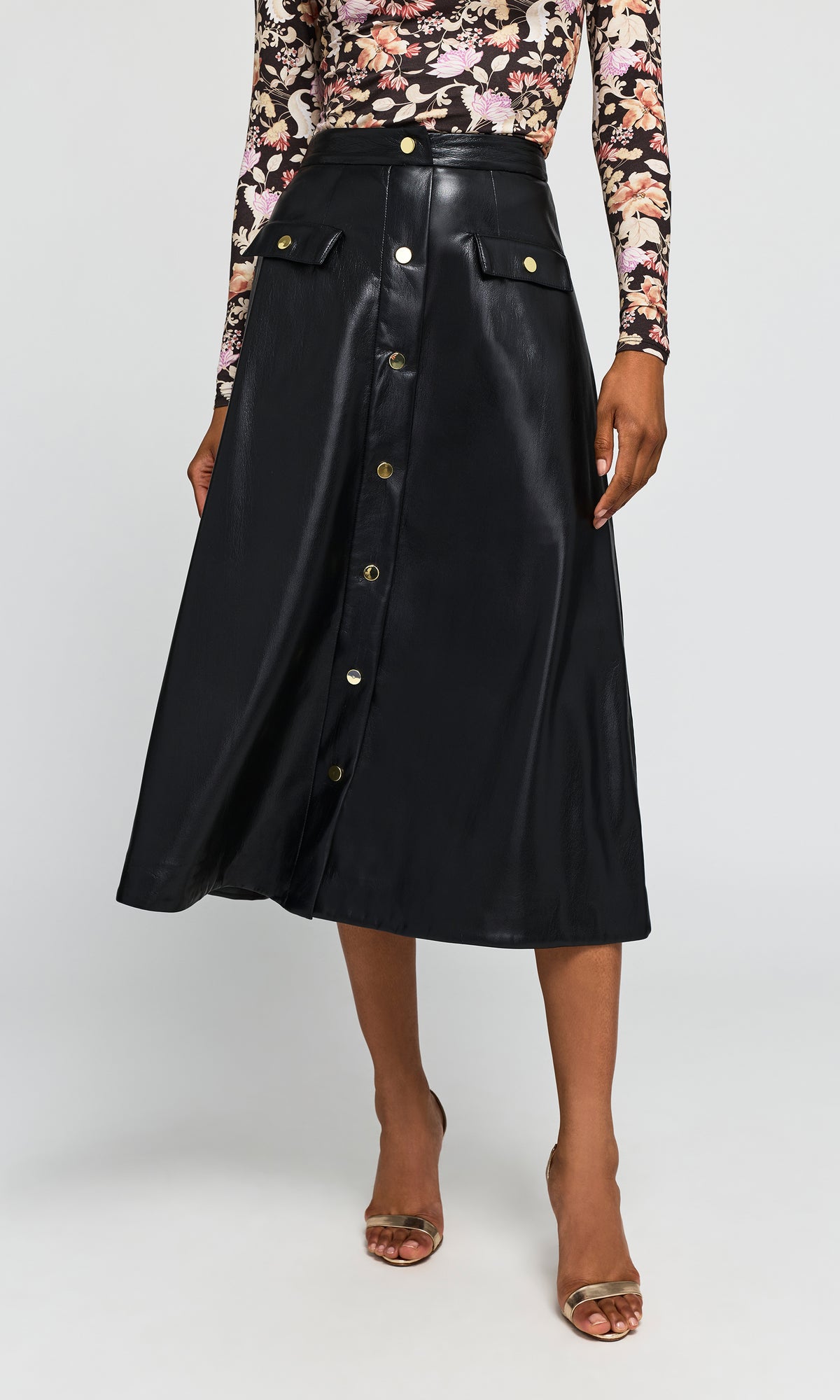 A model wearing a black faux-leather midi skirt with gold-tone button details and flap pockets, paired with a floral long-sleeve top.