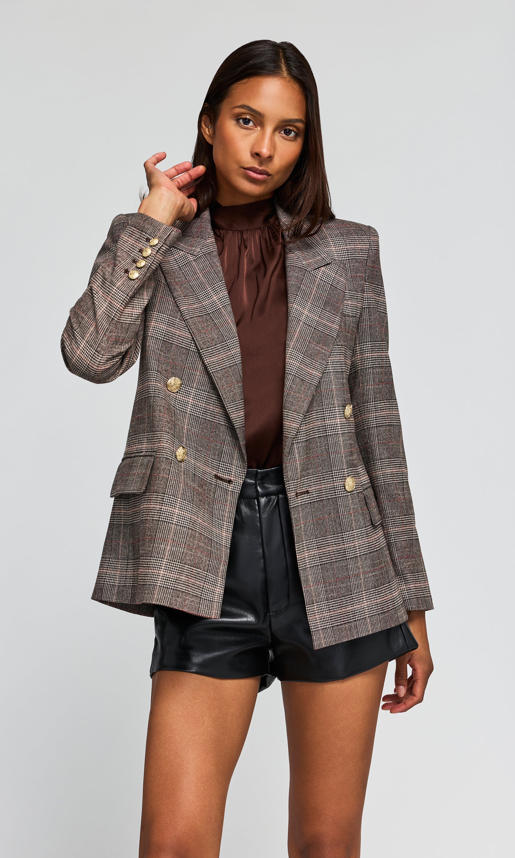 Model wearing a double-breasted plaid blazer in brown and gray tones, paired with a dark brown ruffled top and black shorts.