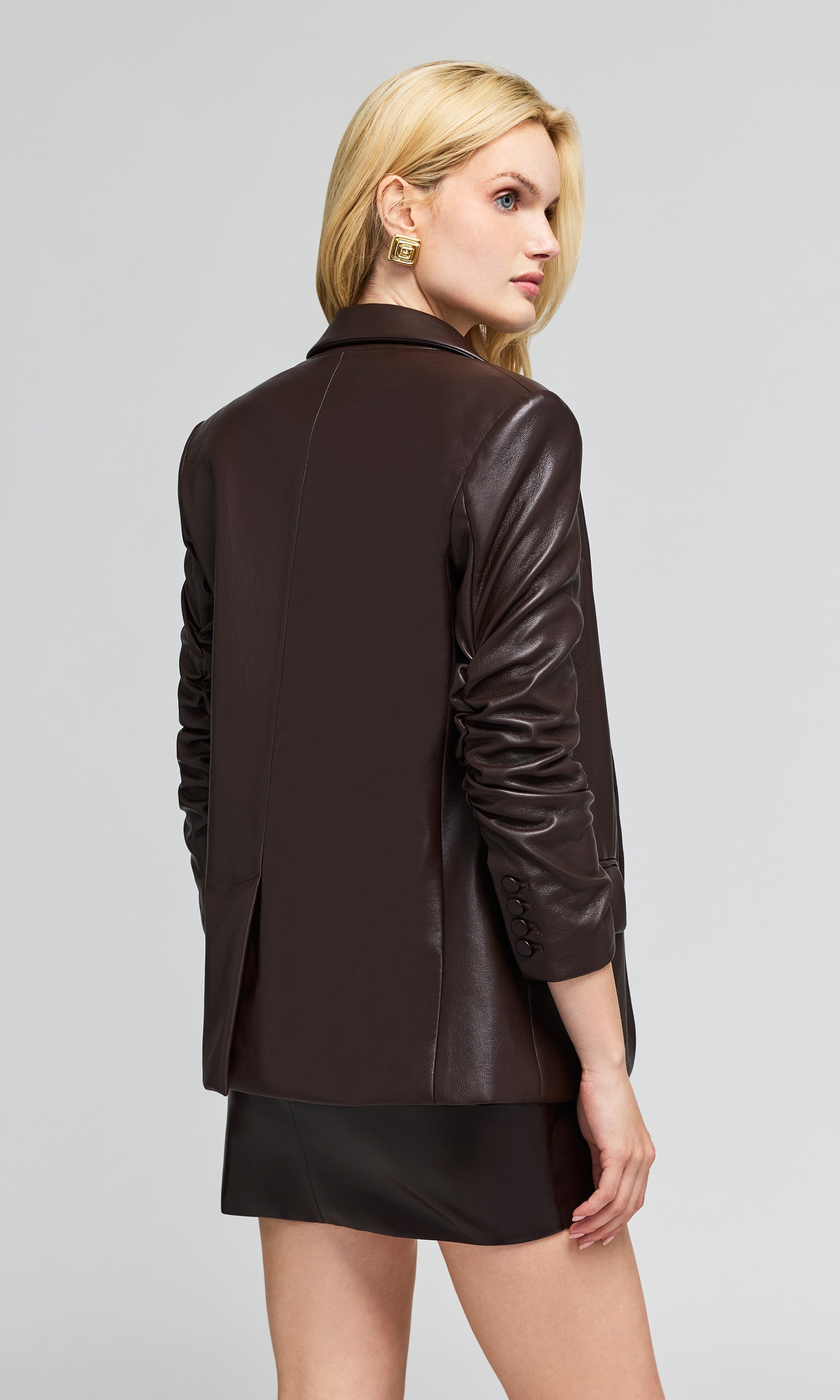 Back view of model wearing a dark brown faux-leather blazer and matching mini skirt on a plain background.