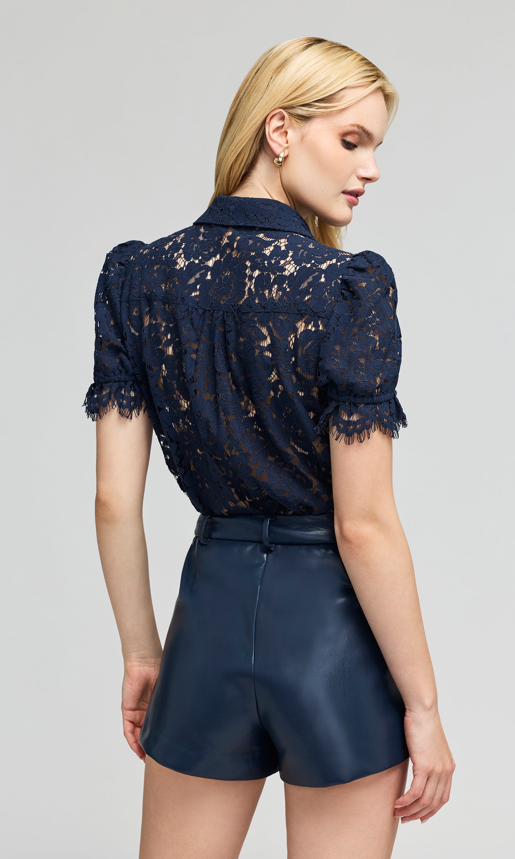 Back view of model shown from the back wearing a navy blue lace blouse dark navy faux-leather shorts, on a plain background.