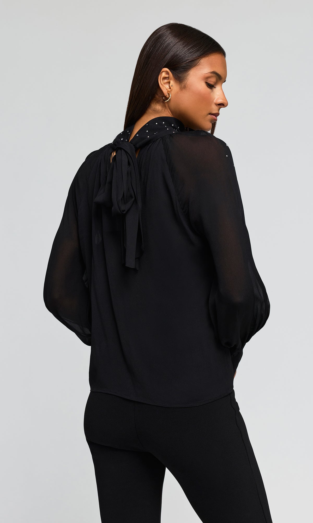 Back view of model wearing a black sheer long-sleeve blouse with a tie-back detail and small silver embellishments.