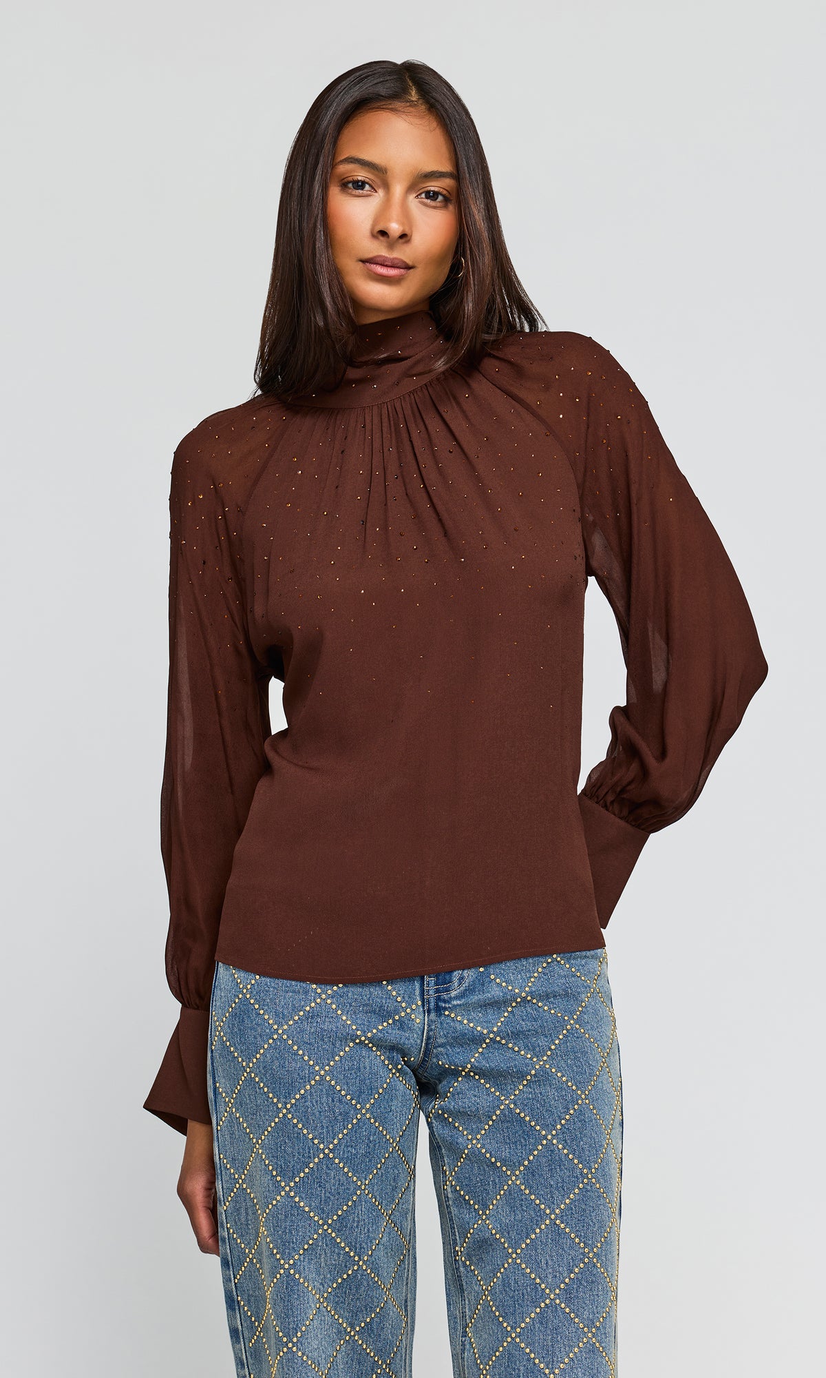 Model wearing a brown long-sleeve blouse with a high gathered neckline and subtle crystal embellishments, paired with blue jeans.