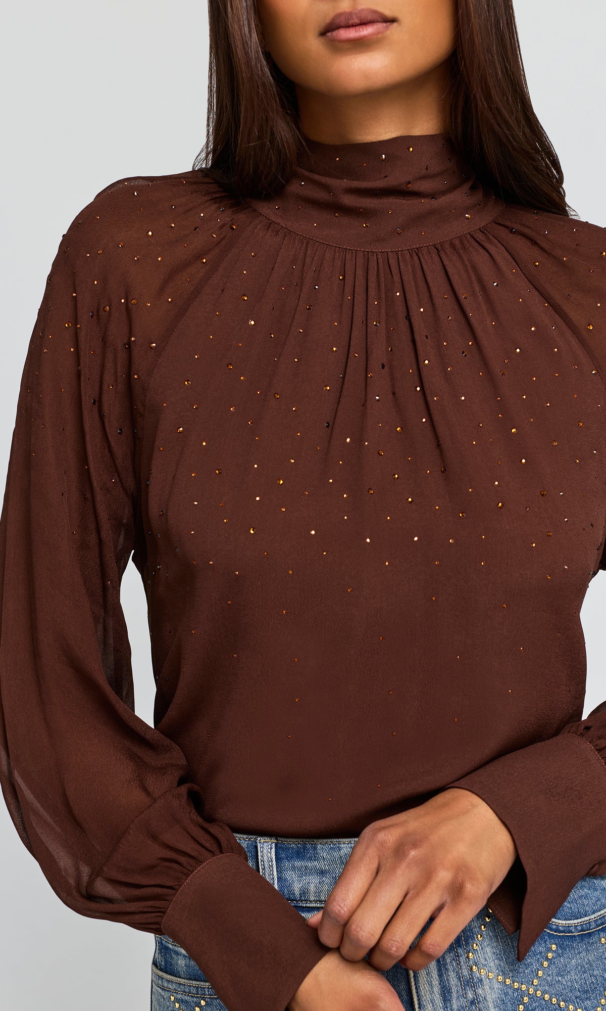 Close-up of a brown long-sleeve mock neck blouse with sheer puff sleeves and small gold sequin embellishments, paired with blue jeans.