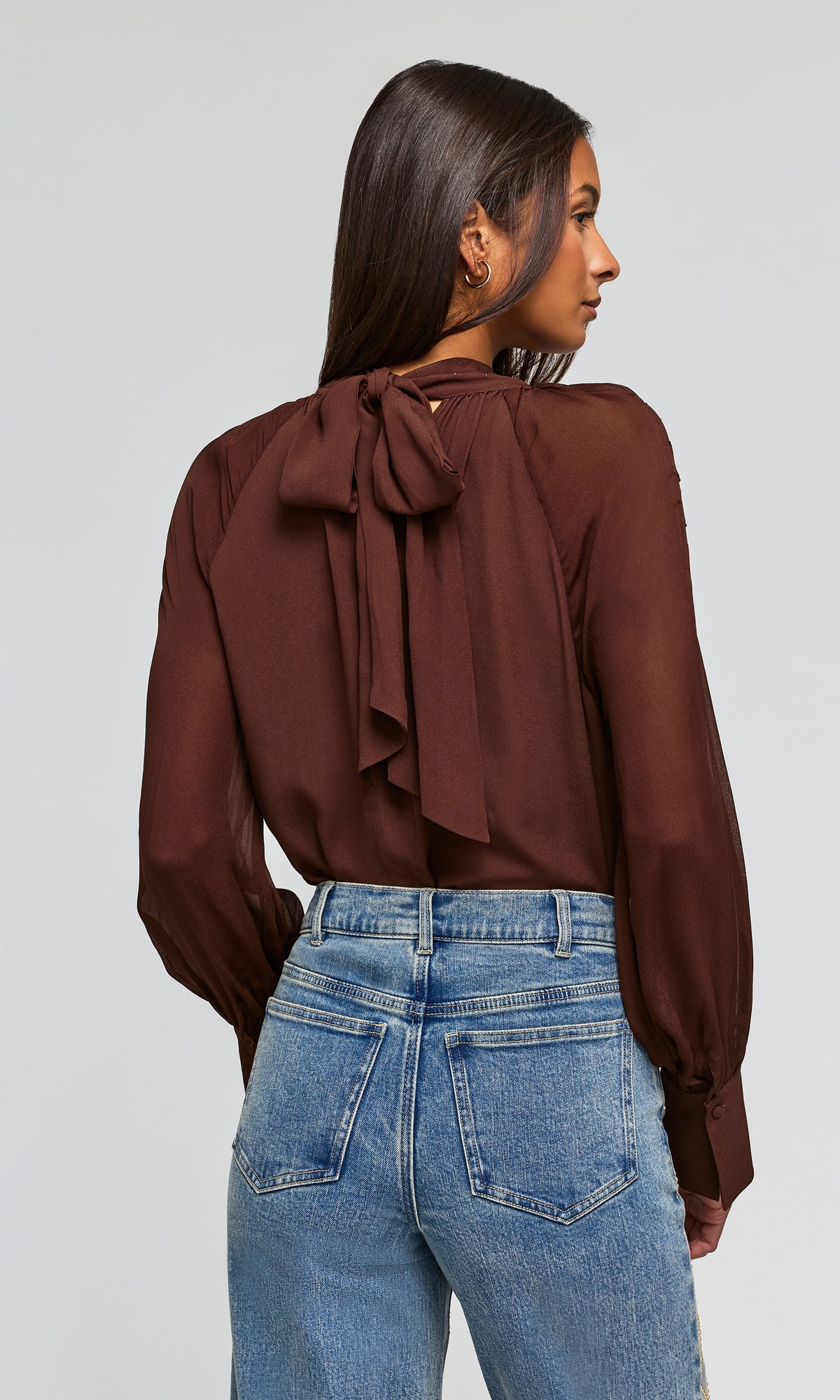 Back view of model wearing a brown long-sleeve blouse with a large bow detail at the neckline and blue jeans.