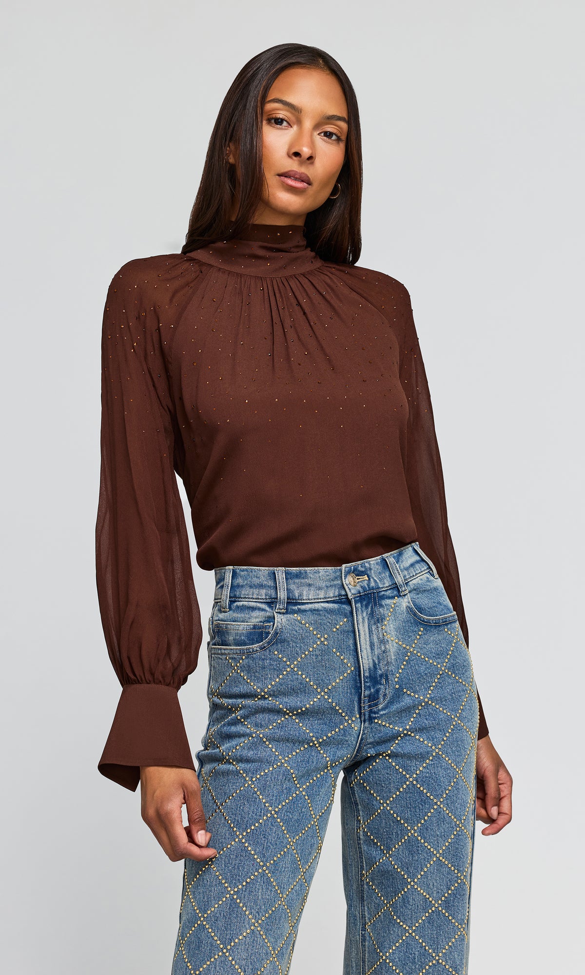 Side view of model wearing a brown long-sleeve blouse with a high gathered neckline and subtle crystal embellishments, paired with blue jeans.