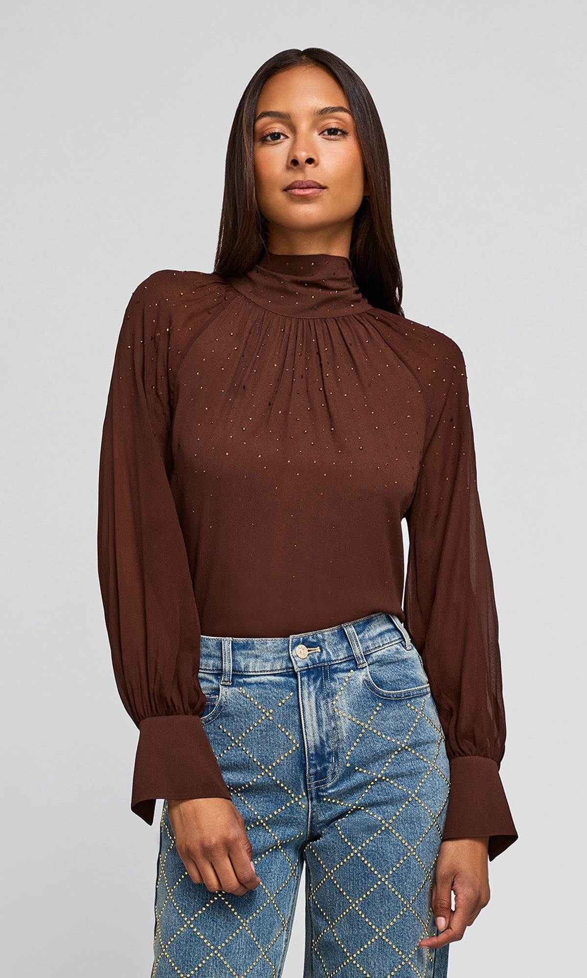 Model wearing a brown long-sleeve blouse with a high gathered neckline and subtle crystal embellishments, paired with blue jeans.