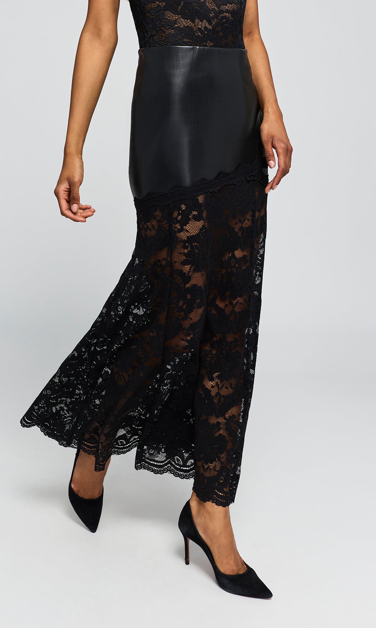 Side view of model wearing a black midi skirt featuring a smooth faux-leather upper section and a sheer black lace lower skirt.