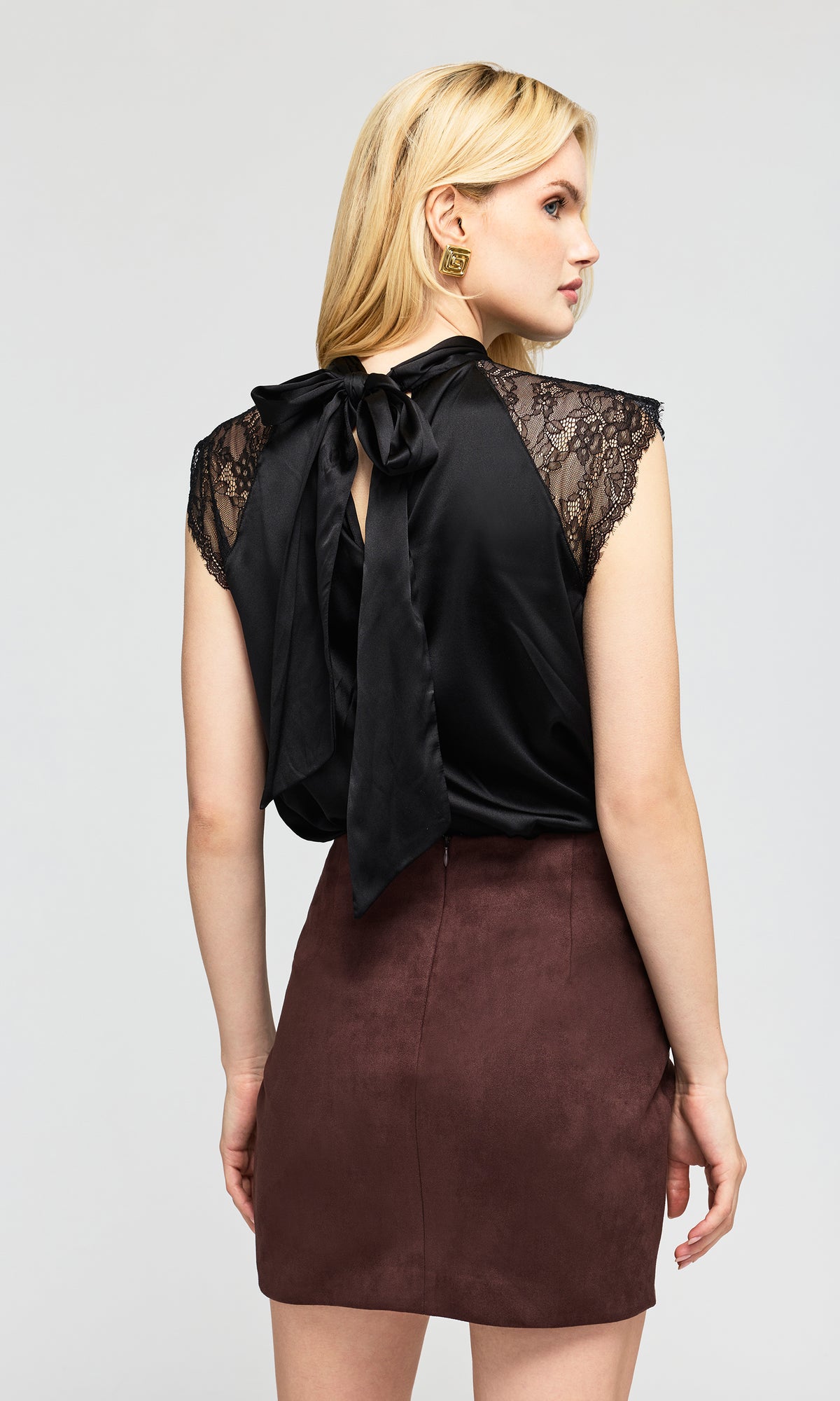 Back view of model wearing a black satin sleeveless top with lace shoulder panels and a large bow, paired with a brown mini skirt.