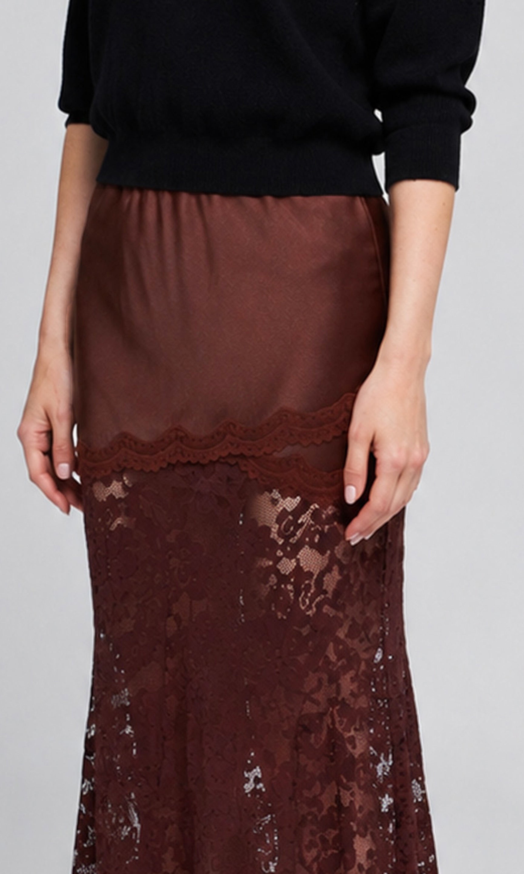 A close-up of a model wearing a dark brown satin skirt with a lace overlay and a scalloped lace hem.