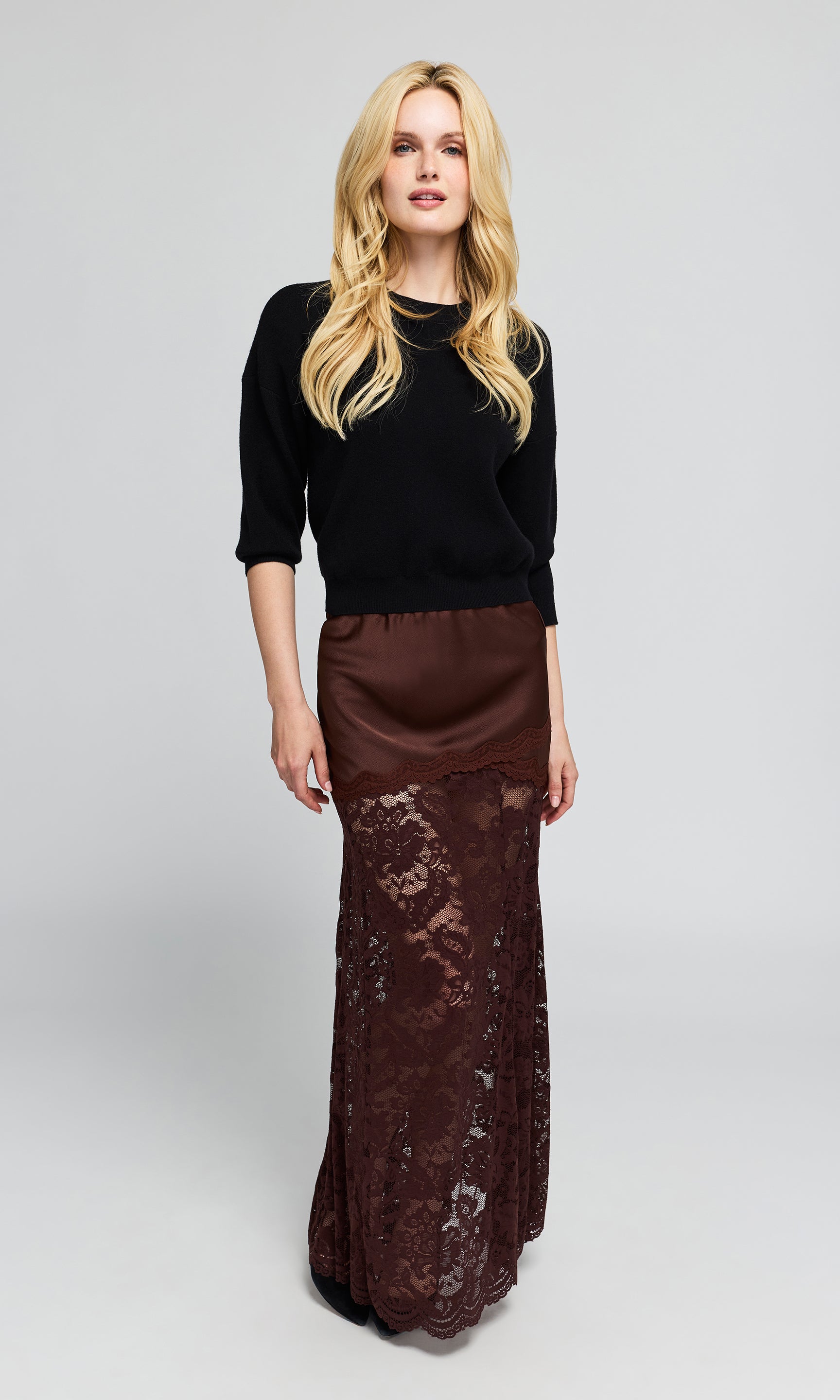 A blonde woman stands wearing a black three-quarter-sleeve top and a long brown skirt.